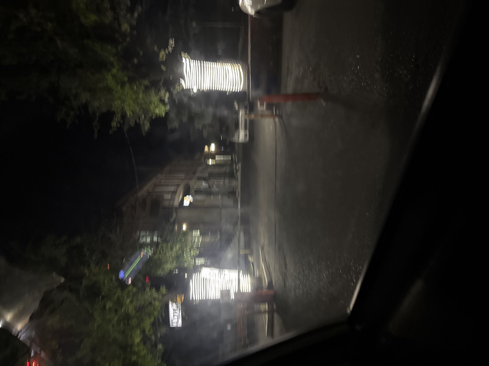 The image depicts a nighttime street scene, likely in Vladikavkaz, Russia, viewed from inside a vehicle. The foreground is dominated by the dark interior of the car, with a sliver of the windshield frame visible on the left. Beyond the car, the street is wet and reflects the surrounding lights. A bright, cylindrical light fixture, wrapped in horizontal bands of light, stands prominently on the right side of the road, casting a strong illumination. A dark, textured barrier or wall runs horizontally across the middle ground, behind the light fixture.
To the left of the light fixture, a wooden bench is visible, accompanied by a dark, vertical object that could be a trash receptacle or a bollard. Further back, several buildings are dimly lit, with some windows appearing bright, suggesting internal lighting. One building on the left has a rectangular sign with blurry, white text. Trees with green foliage are scattered throughout the scene, their leaves illuminated by the artificial lights. In the middle ground, a white van is parked on the street. The overall impression is a quiet, urban street at night, possibly a pedestrian area or a side street with limited activity.