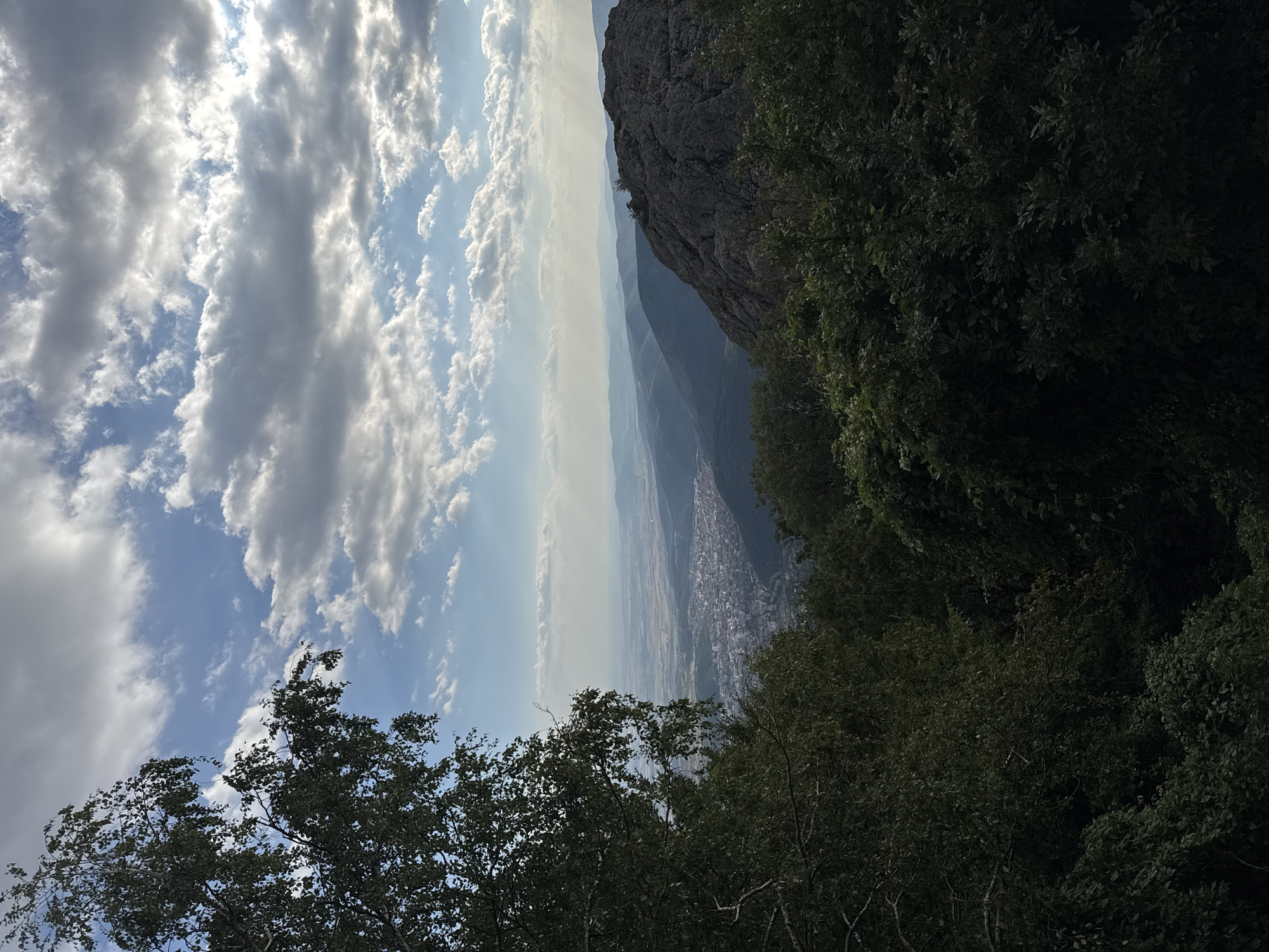 The image is a scenic vista overlooking a city, identified as likely being in the vicinity of Ichera, Bulgaria, given the context. In the foreground, a large tree with dark green foliage frames the left side of the image, its branches extending into the upper left corner. To the right of the foreground, a steep, rocky outcrop with rough texture dominates the mid-ground. Beyond the rocky outcrop, the landscape recedes into a valley where a city is visible, its buildings clustered together and appearing as a grey expanse. Further in the distance, layers of rolling hills fade into a hazy horizon under a dynamic sky. The sky is a mix of blue with scattered white and grey clouds, with shafts of sunlight breaking through in places, suggesting late afternoon or early morning light. The overall impression is one of a natural overlook providing a panoramic view of a populated area amidst a mountainous or hilly terrain. There are no people or discernible actions depicted in the image.