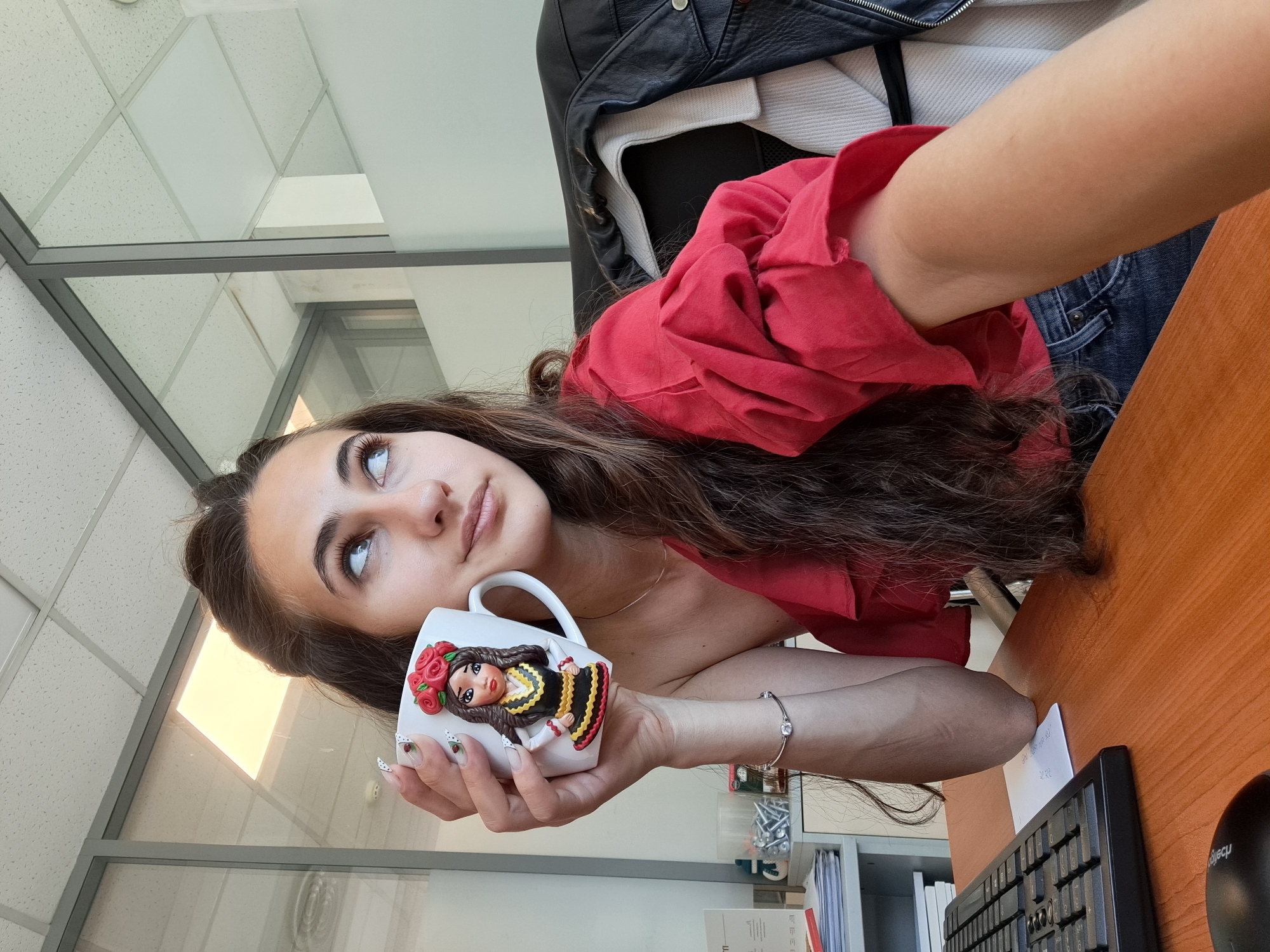 A woman sits at a desk in an office, holding a mug. She is fair-skinned with long brown hair and is wearing a red top. She is looking up and to the left, away from the camera. The mug she holds is white with a 3D figure of a woman dressed in traditional Bulgarian clothing with red roses in her hair. The desk is made of brown wood, and a black keyboard and mouse are on it. There is a window with a gray frame behind her, with a view of what appears to be another office building, which has a ceiling comprised of small white tiles. A black leather jacket is hanging over the chair. There are papers and office supplies visible behind her on a shelf. Given the traditional Bulgarian clothing depicted on the mug, and without explicit confirmation, it is possible the photo was taken in Sofia, Bulgaria, but that is not definitive from the image alone.