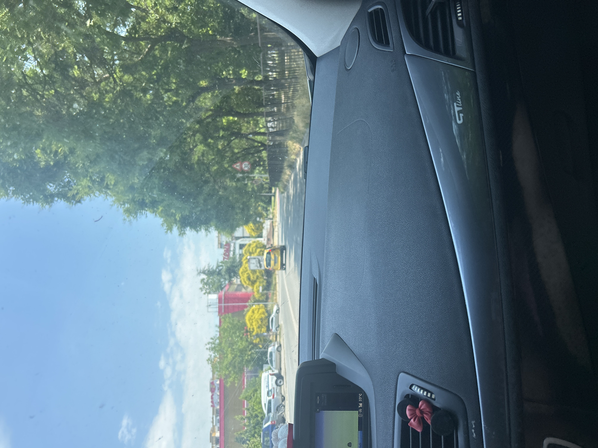 The image is taken from the interior of a car, looking forward through the windshield. The car is in motion, driving down a street in Plovdiv, Bulgaria, on a sunny day. The dashboard is visible in the foreground, with a navigation screen displaying a map and the temperature as 33°C. There is a car air freshener attached to the dashboard. Outside the car, there are trees lining the street and buildings in the background, including a red building with signs. Several cars are parked on the side of the road, and a yellow car is driving ahead. A speed limit sign (30) is visible. The overall environment suggests a suburban or commercial area within the city.