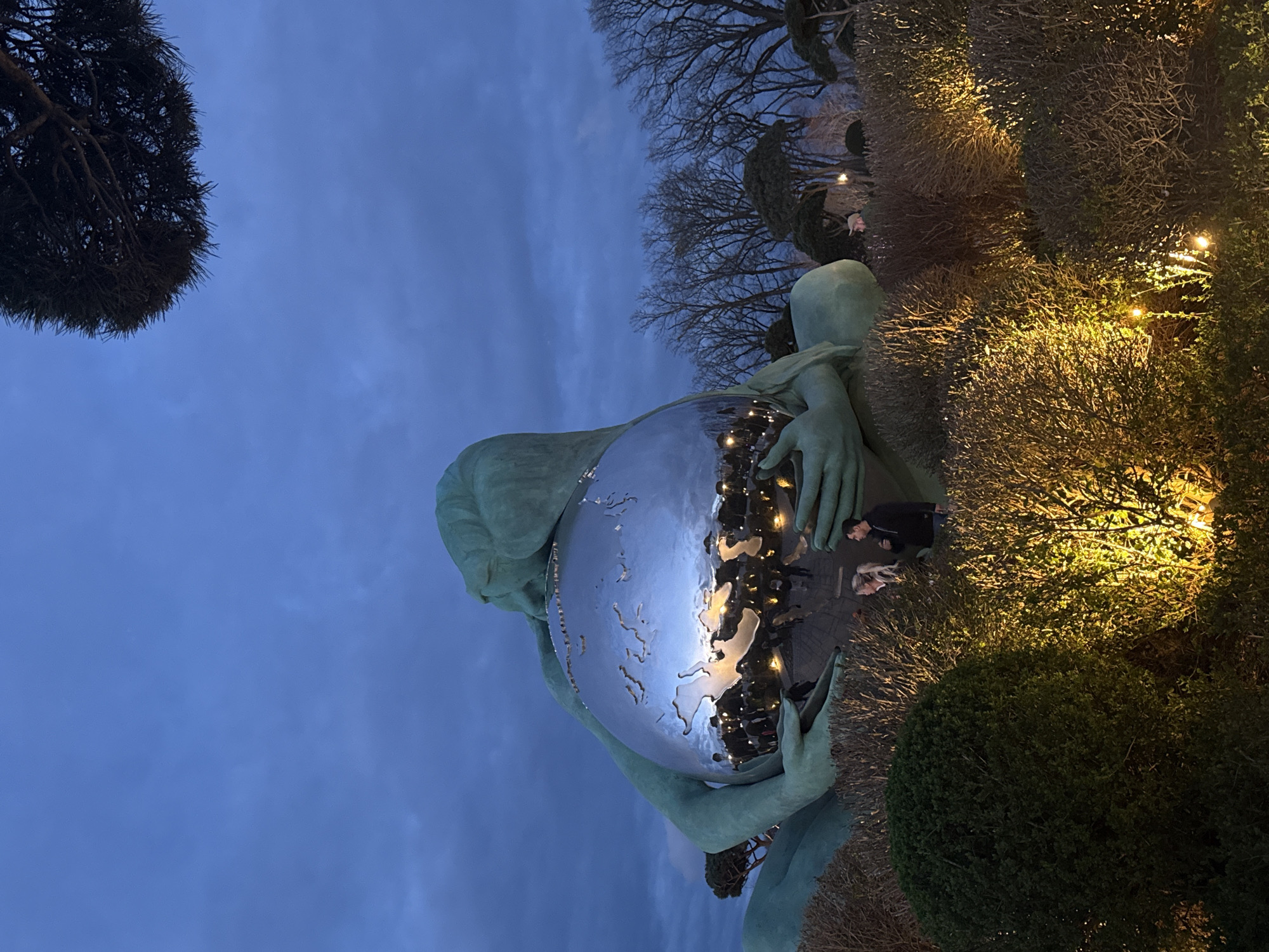 A low-angle shot depicts a statue of a figure holding a shiny, spherical representation of the world. The statue is situated among bushes and trees, some adorned with small, warm-toned lights. Two individuals, one with blond hair, are visible among the bushes near the base of the statue. The sky behind the statue is a deep blue, suggesting it is dusk or evening. The reflection in the sphere shows a city scene with buildings and street lights. It is located in the city of Unknown, Unknown.