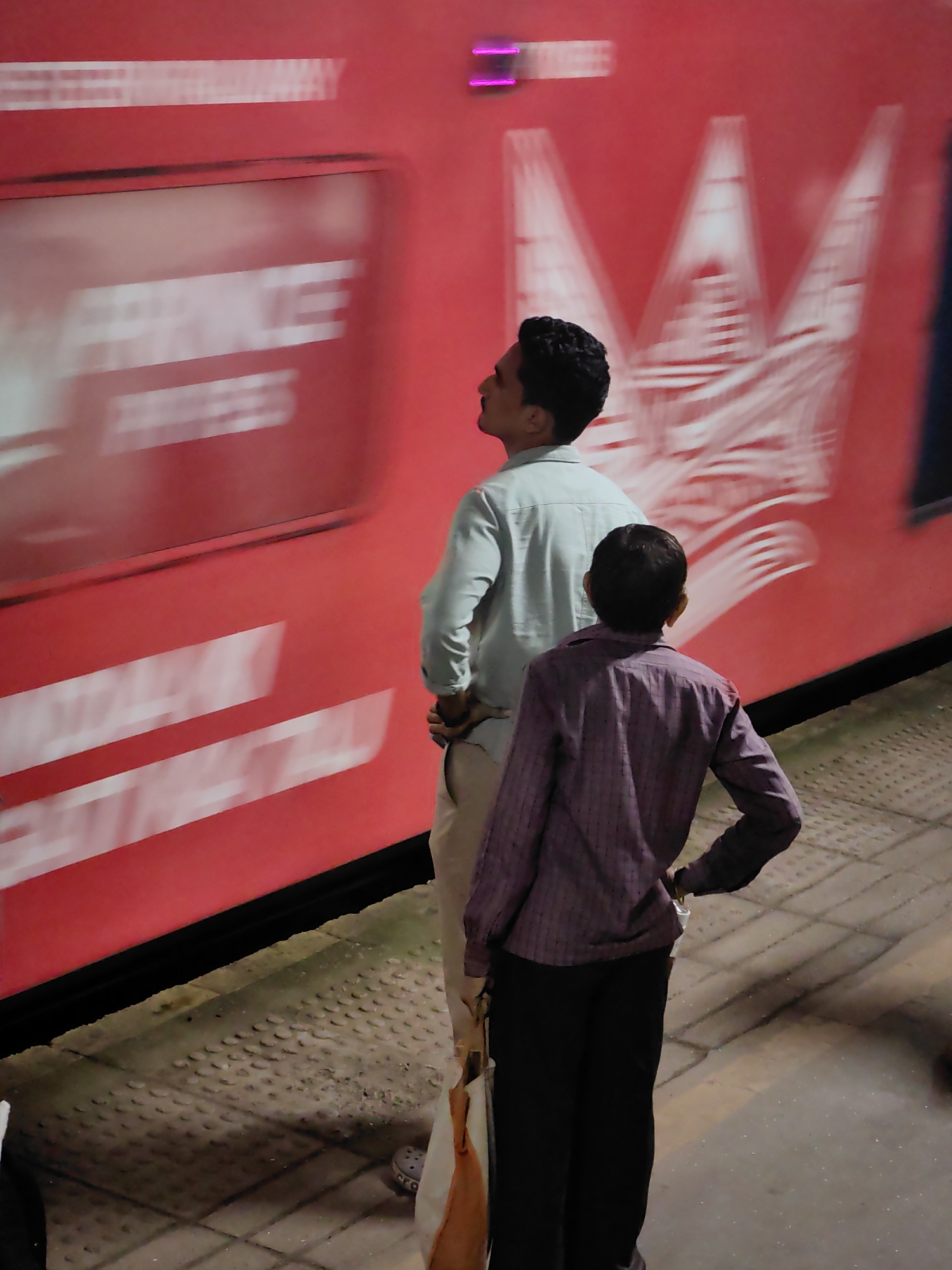 The image depicts two individuals standing on a platform next to a red train. The train appears to be in motion, as evidenced by motion blur on its surface and the text visible on its side. The person on the left is wearing a light-colored shirt and light-colored pants, looking up at the train. The person on the right is wearing a purple checkered shirt and black pants, also looking towards the train. The platform is paved with textured tiles, characteristic of Indian railway stations. A bag is visible next to the person on the right. The environment suggests a railway station in Mira Bhayandar, India.