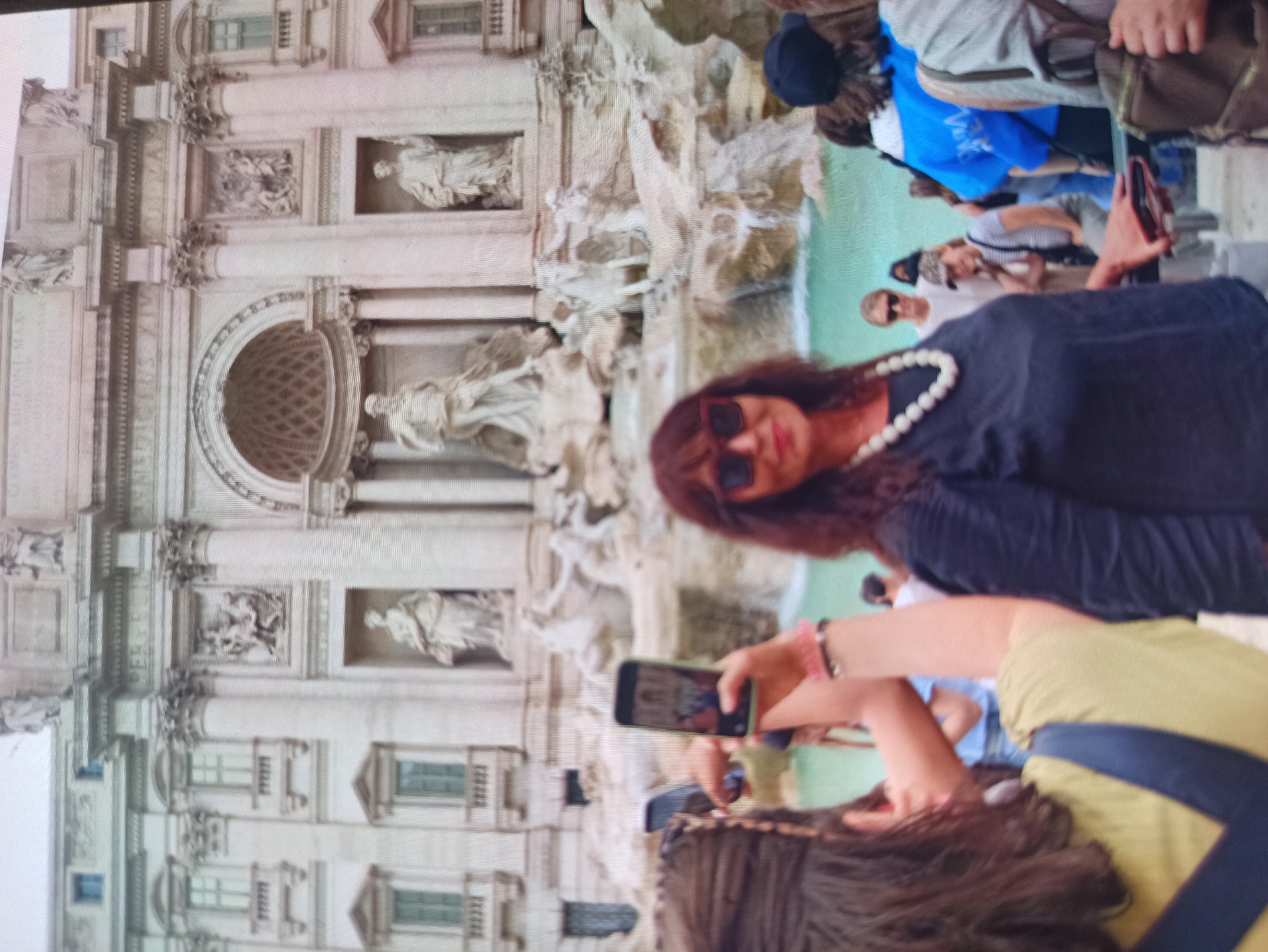 The image depicts the Trevi Fountain in Rome, Italy, a famous landmark. In the foreground, several people are visible, many holding up their phones to take pictures or videos. A woman in the center foreground is wearing a dark top and a pearl necklace, and she is holding a smartphone. To her left, another person with dark hair is holding a phone, taking a picture. The background shows the elaborate Baroque architecture of the fountain, with statues, columns, and cascading water. The overall scene suggests a tourist attraction with many visitors. The image does not provide any information or context related to Kryvyi Rih, Ukraine.