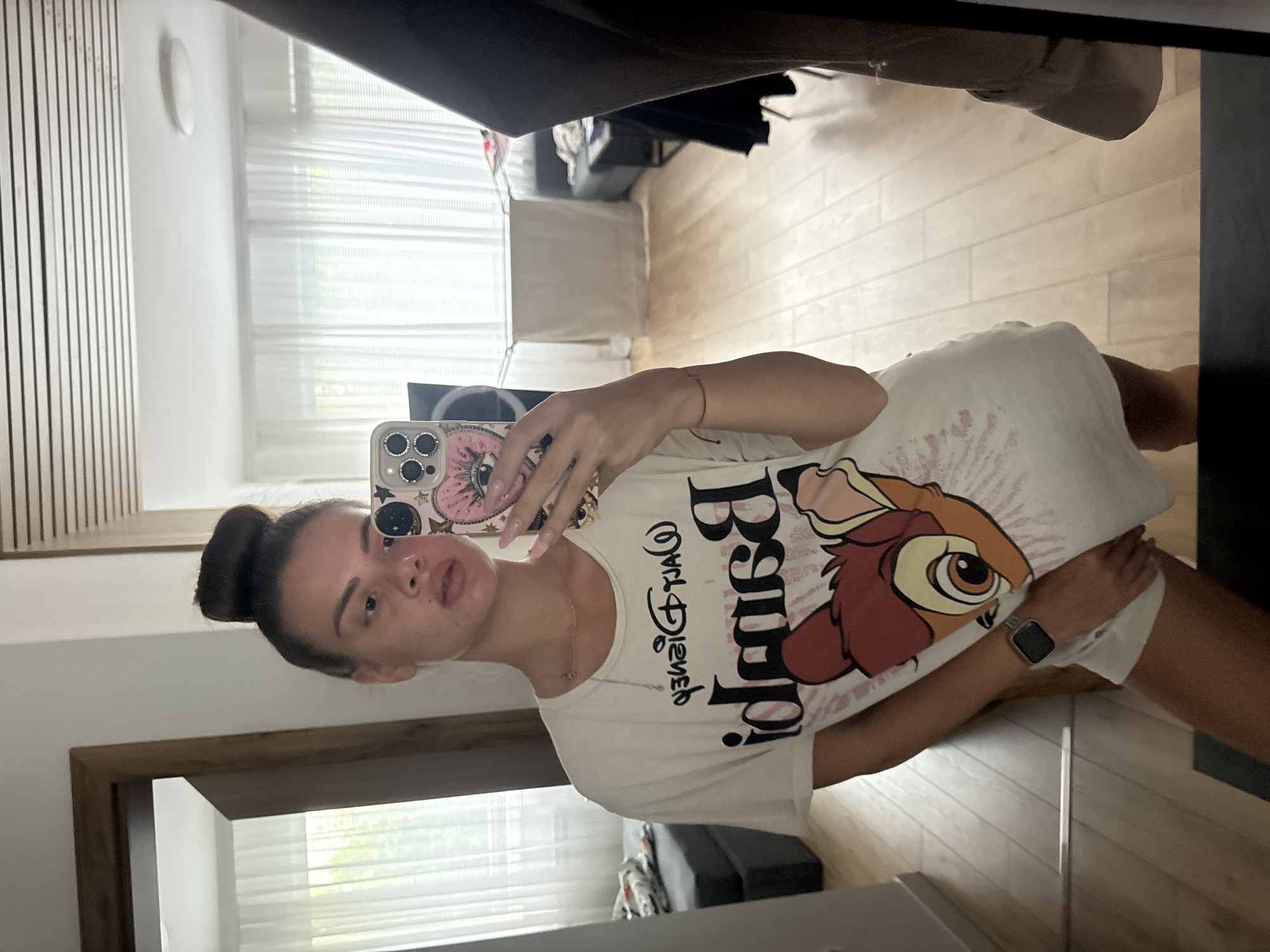 A young woman with her hair in a top knot, wearing an oversized white t-shirt with a Bambi graphic, stands in front of a mirror in what appears to be a bedroom. She is taking a selfie with her phone, which has a pink case decorated with eyes and stars. She is wearing a silver watch on her left wrist and a thin red bracelet. The room has wooden floors and large windows covered by sheer white curtains. In the background, a dark couch is visible. The scene is set in Plovdiv, Bulgaria.