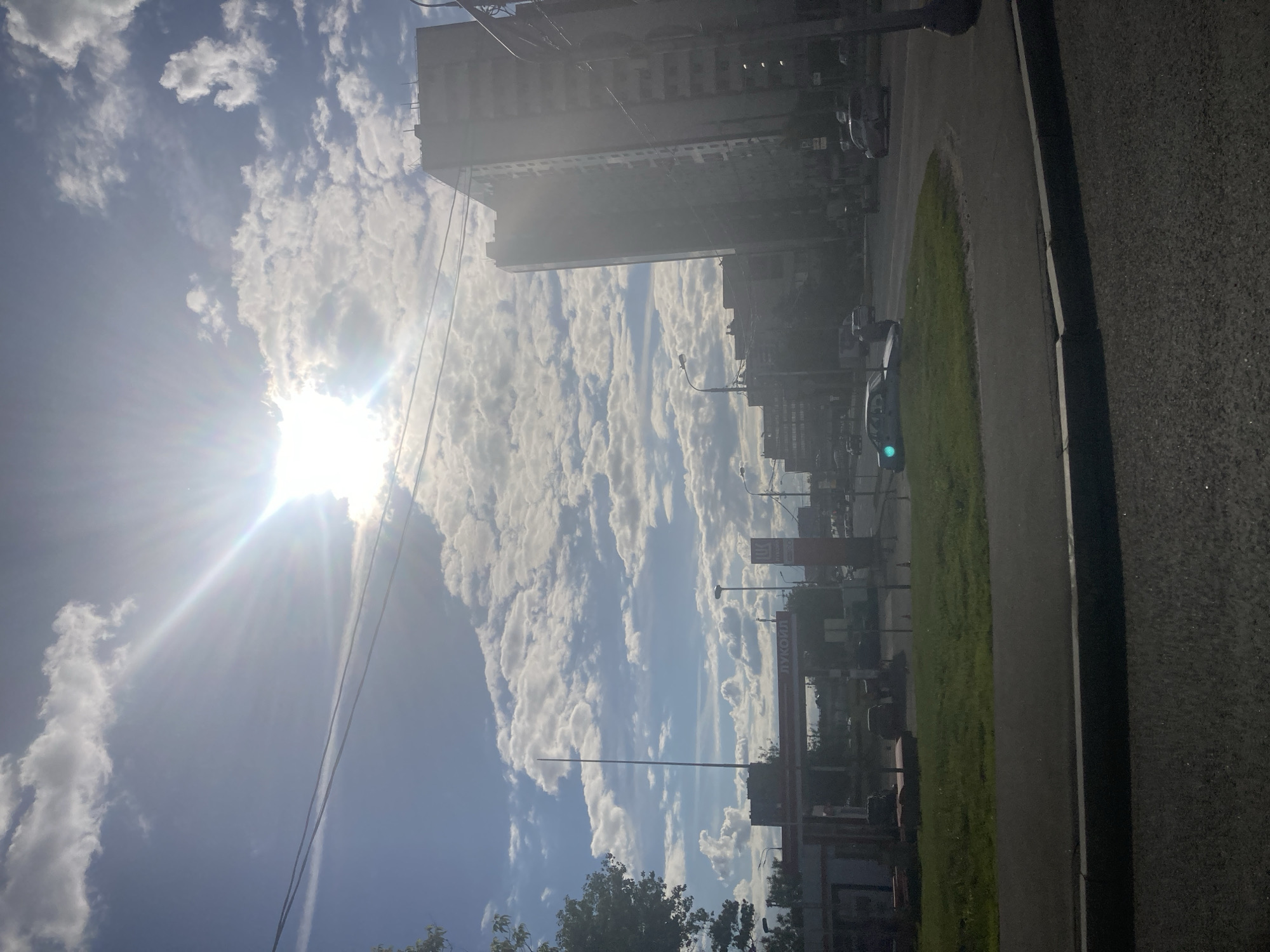 The image shows a street scene in Khimki, Russia, taken on a sunny day. A tall building is visible in the background. The sky is filled with cumulus clouds. A Lukoil gas station can be seen on the right side of the image. There is a grassy area between the sidewalk and the street. A car is stopped at a green traffic light. Electrical wires cross the sky.