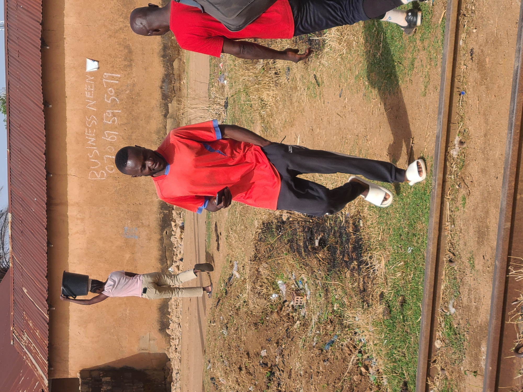 The image depicts an outdoor scene in Jos, Nigeria. Two men are prominently featured: one wears a red shirt with blue trim, dark trousers, and white sandals. He holds a dark object, possibly a phone. The other man, further in the background, wears a red shirt, dark trousers, and sneakers, and carries a dark bag. Another person in light-colored clothing is visible in the distance.
The scene is situated near what appears to be a building with a rusty metal roof. Written on the wall of the building is "BUSINESS NEEN" followed by the phone number "07067595079." The ground is a mix of dirt, grass, and scattered trash. A set of railroad tracks runs through the area.