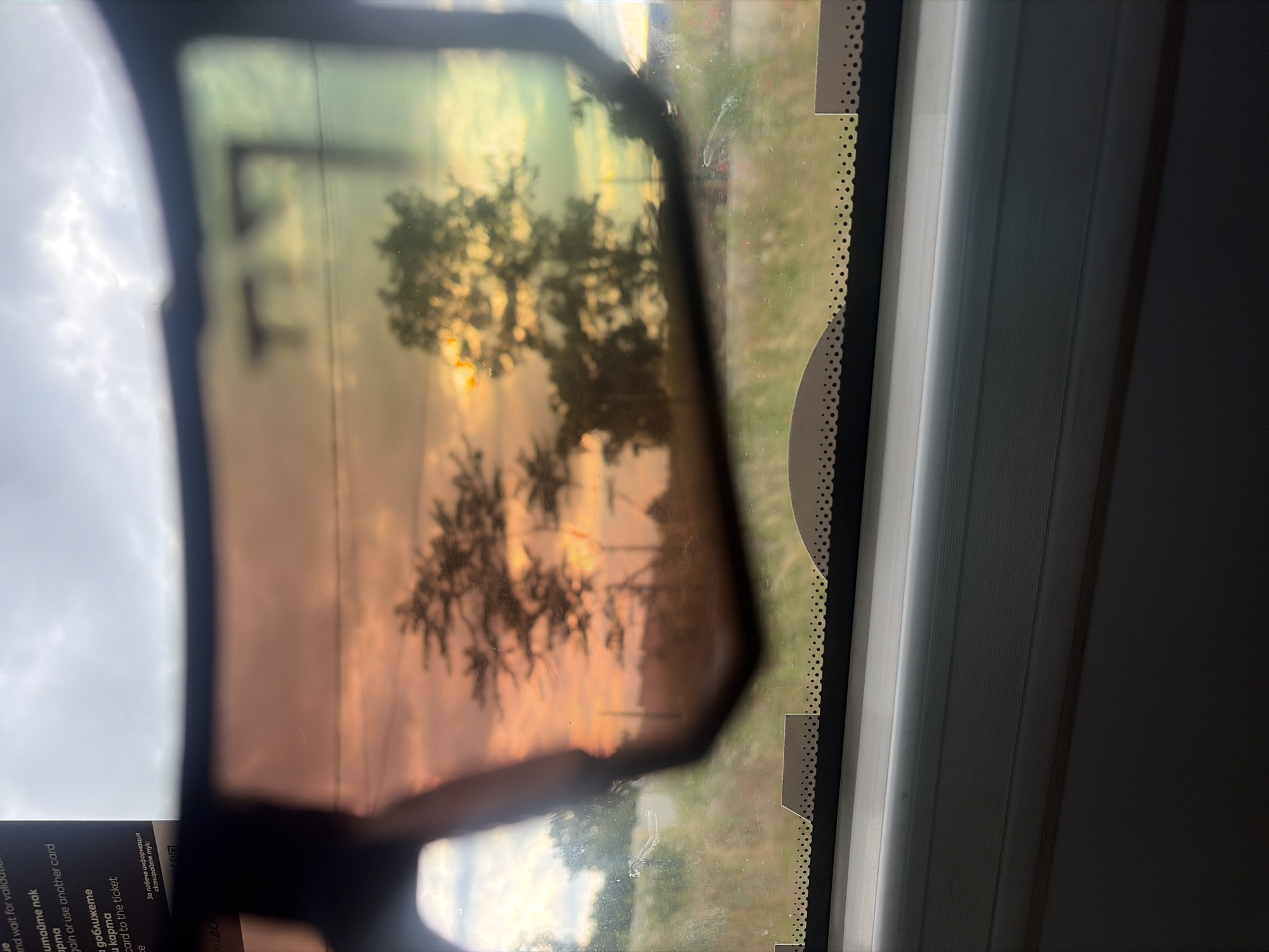 The image shows a view through a window with a pair of sunglasses in the foreground, casting a warm, colored tint on the scene. The sunglasses are partially obscuring a view of trees silhouetted against a sky at either sunrise or sunset, with colors ranging from orange and yellow to green. The environment outside appears to be rural or semi-rural, with fields and vegetation visible. The interior of the vehicle, possibly a train or bus, is visible at the bottom of the frame, with a window ledge and possibly blinds. A card with some text is also partially visible in the upper left corner. No people are visible in the image. The location of Kubratovo, Bulgaria is not discernible from the image itself.