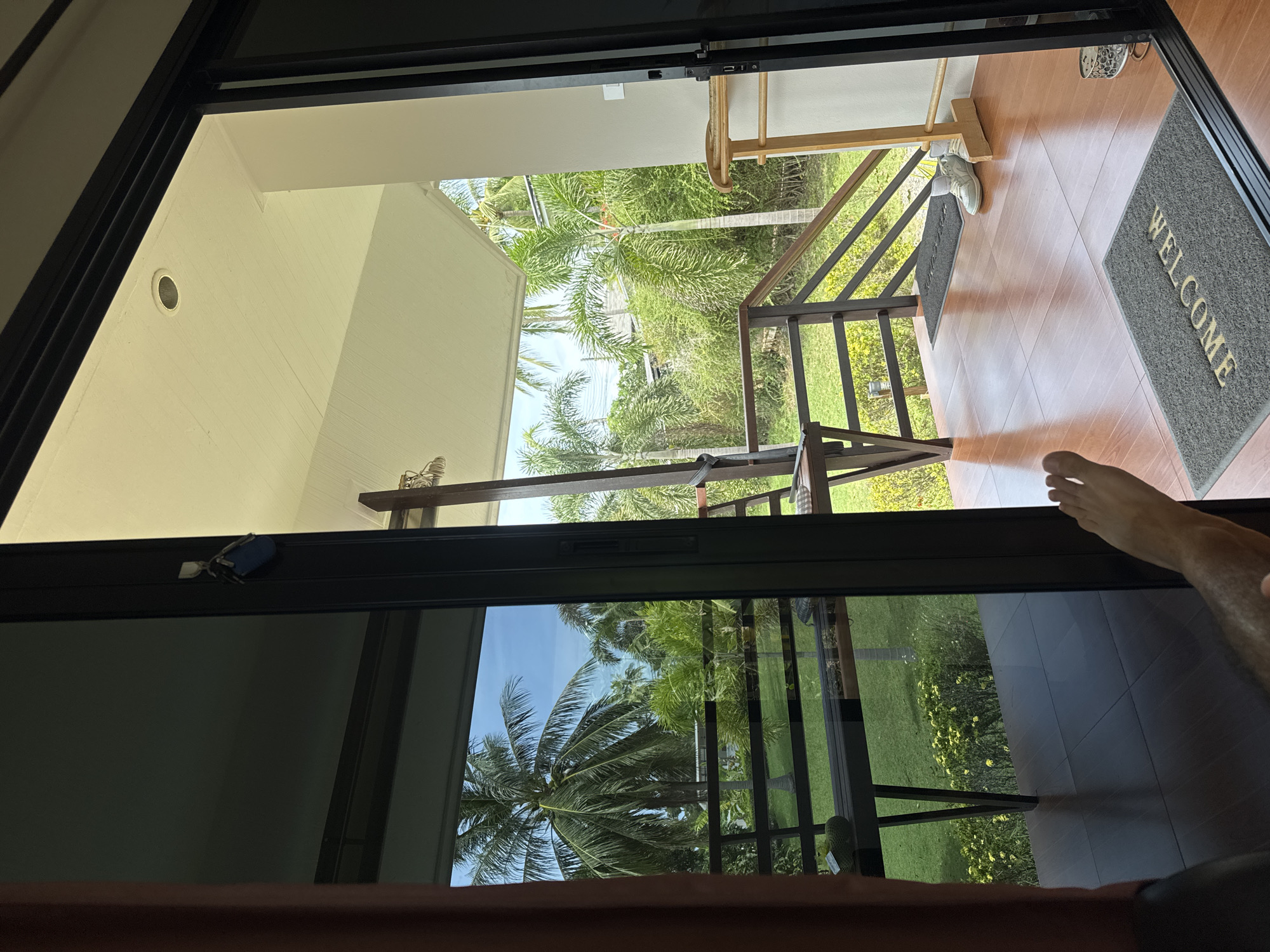 The image shows a view from inside a room looking out onto a covered patio and tropical garden. The patio has a wooden floor and a railing overlooking a grassy area with lush green vegetation and palm trees. A pair of white sneakers and a wooden coat rack are visible on the patio. A doormat with the word "WELCOME" is placed near the entrance. In the reflection of the glass door, a person's foot and leg are visible, suggesting they are inside the room. The scene evokes a sense of tropical relaxation, possibly in a resort or villa in Ko Samui District, Thailand.