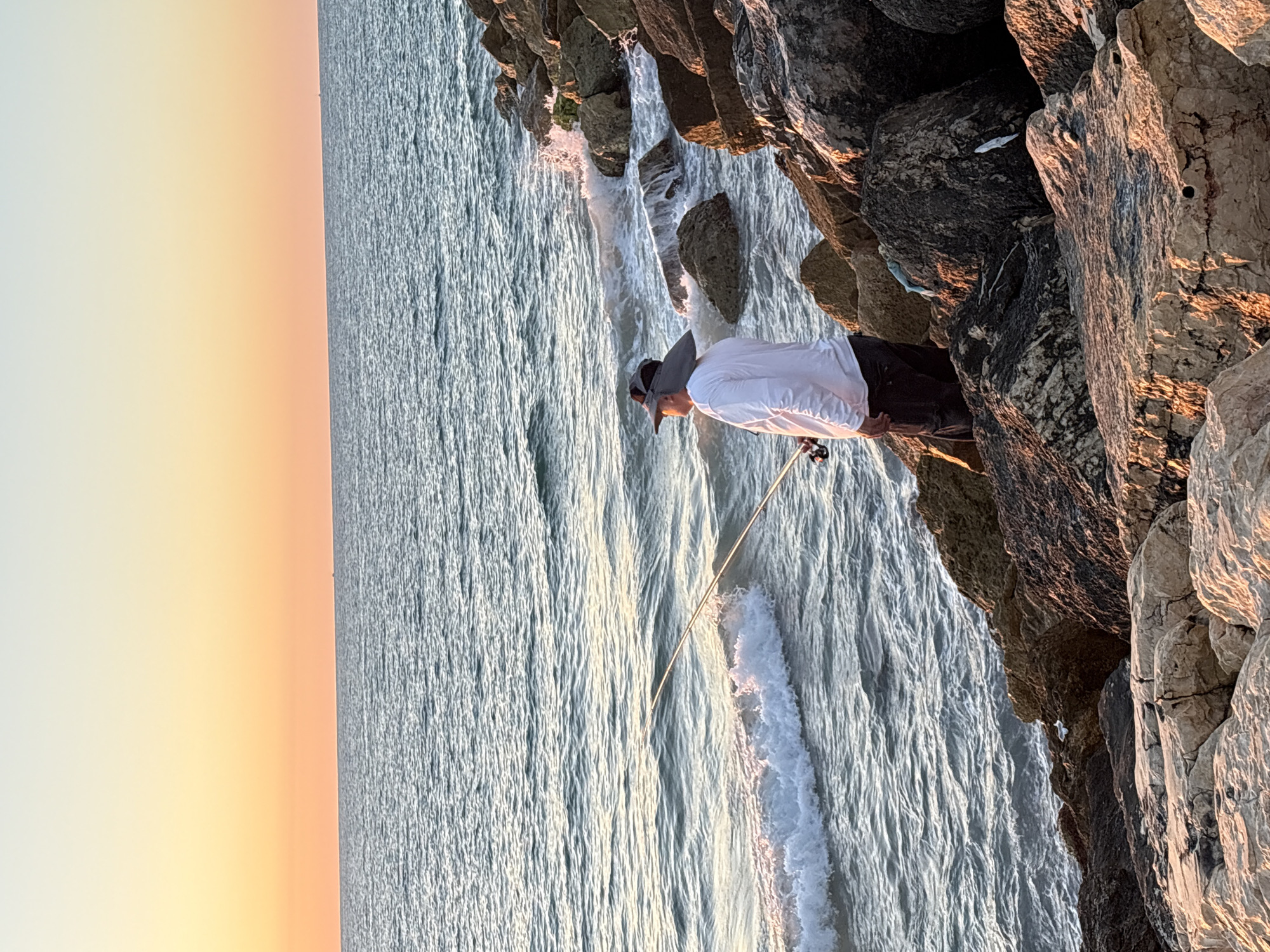 A lone fisherman stands on a rocky shore, casting a line into the gentle waves of the Mediterranean Sea at sunset. The sky displays a gradient of soft orange and yellow hues, characteristic of a Tel-Aviv sunset. The fisherman, wearing a wide-brimmed hat and a white long-sleeved shirt, is focused on the water, his fishing rod held steady. The rocks, weathered by the sea, form a rugged embankment. The ocean's surface reflects the warm colors of the sky, and small waves break against the shore. The overall scene evokes a sense of peaceful solitude.