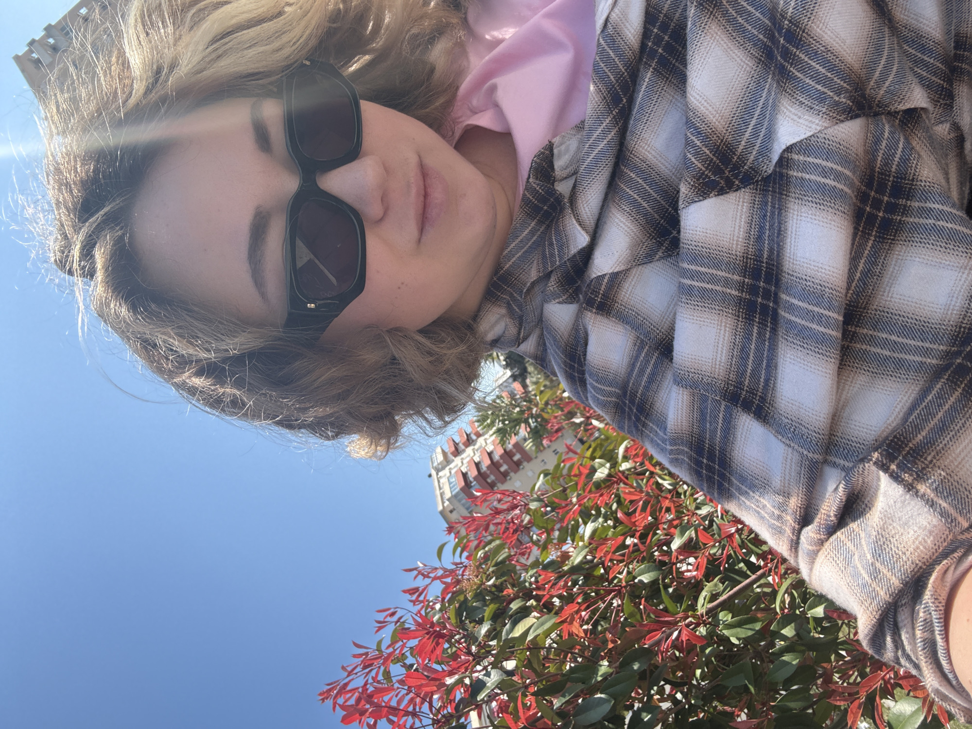 A selfie taken from a low angle shows a person with blonde, wavy hair wearing dark sunglasses, a pink hoodie, and a plaid shirt. The sky is clear and blue. In the background is foliage with red-tinged leaves. A multi-story building with what appears to be red window coverings is visible in the upper center of the frame. Given the city mentioned, this is likely in Bar, Montenegro, although specific landmarks are not discernible. The person is likely outdoors, with the foliage suggesting a park or streetscape.