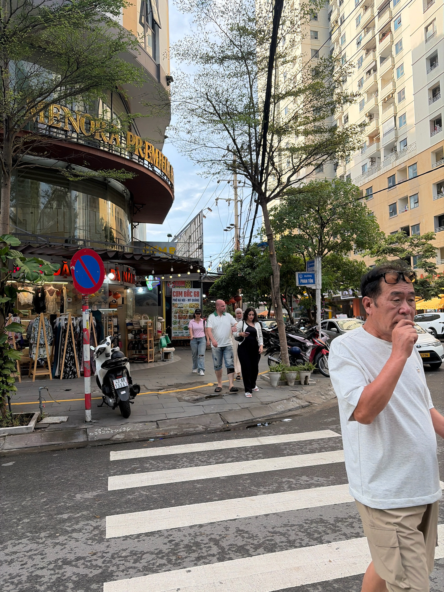 A man in a white t-shirt and tan cargo shorts stands on a crosswalk in the city of Da Nang, Vietnam. He is looking to his right and has sunglasses perched on his head. Behind him, several people are walking on a sidewalk in front of shops. A white scooter is parked on the sidewalk. Buildings line the street, including a tall apartment building. The sky is overcast.