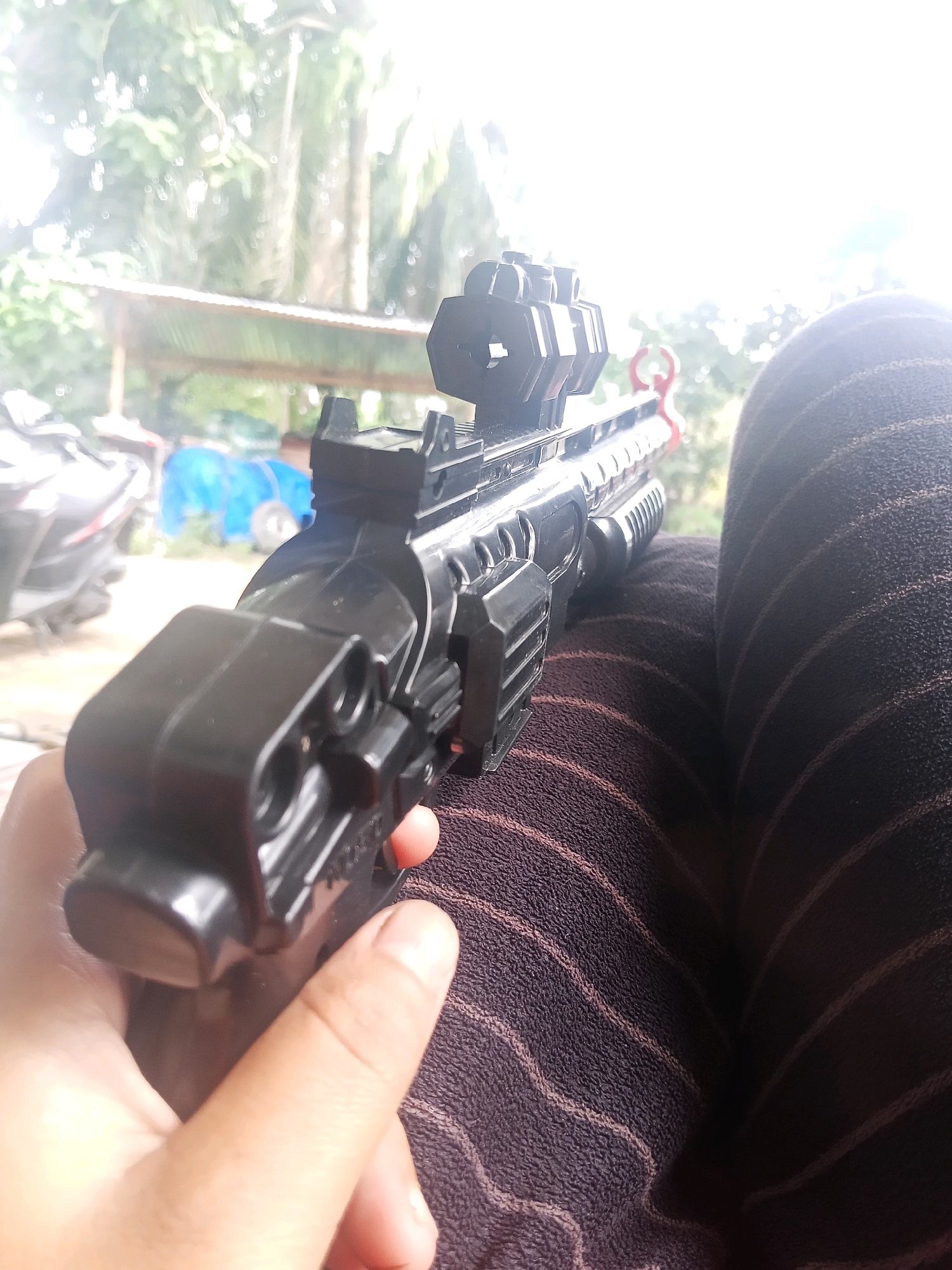 A close-up, slightly blurry, high-angle shot shows a black toy gun with a scope being held by a person's hand. The toy gun is the primary subject, with its textured grip and scope clearly visible. The person's hand is a lighter skin tone, and a portion of their legs, clad in dark striped pants, is also in the frame. The background is out of focus, revealing a blurred outdoor scene with greenery and what appears to be a structure with a corrugated roof. There are also hints of what might be motorcycles and a blue tarp in the distance, suggesting a residential or semi-rural environment. The lighting is bright, with a significant portion of the upper right of the image being overexposed, possibly due to sunlight. There is no clear indication of the city of Lestari Indah, Indonesia, in the visual elements of the image.