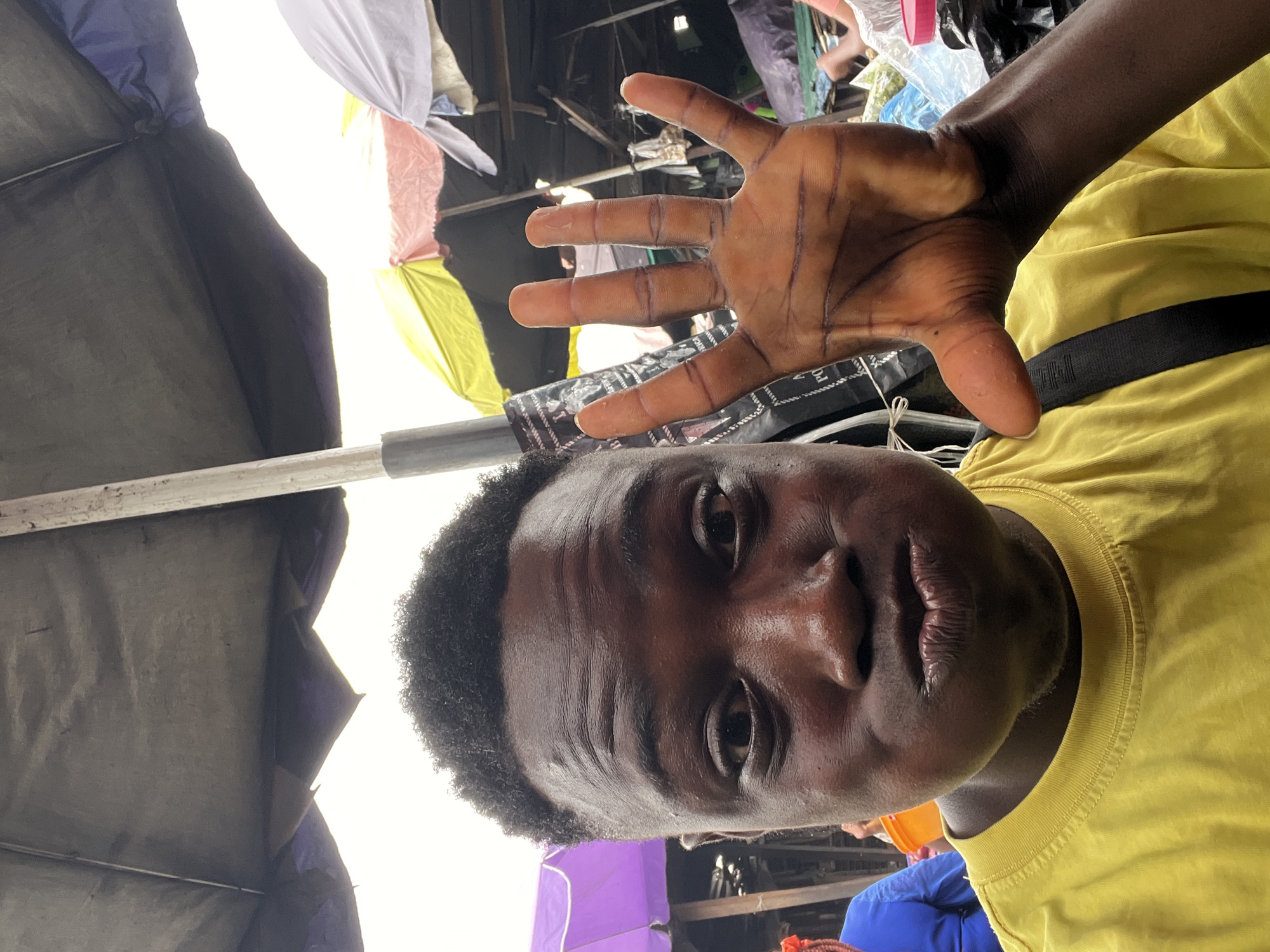 A medium-angle, close-up shot shows a Black male with short, curly hair, wearing a yellow t-shirt with a black strap across his chest, positioned slightly off-center. The male is holding up his hand in front of his face, palm facing forward. The background appears to be a bustling market scene, possibly in Nkpor, Nigeria, as suggested by the image prompt. A partially visible structure above, possibly a market stall, includes a mix of colored fabrics, including pink, white, yellow, and dark-colored tarpaulin. Other figures are blurred in the distance, and hints of clothing like blue fabric suggest a busy environment. The lighting appears to be bright and overcast, with a focus on the subject's face and hand.
