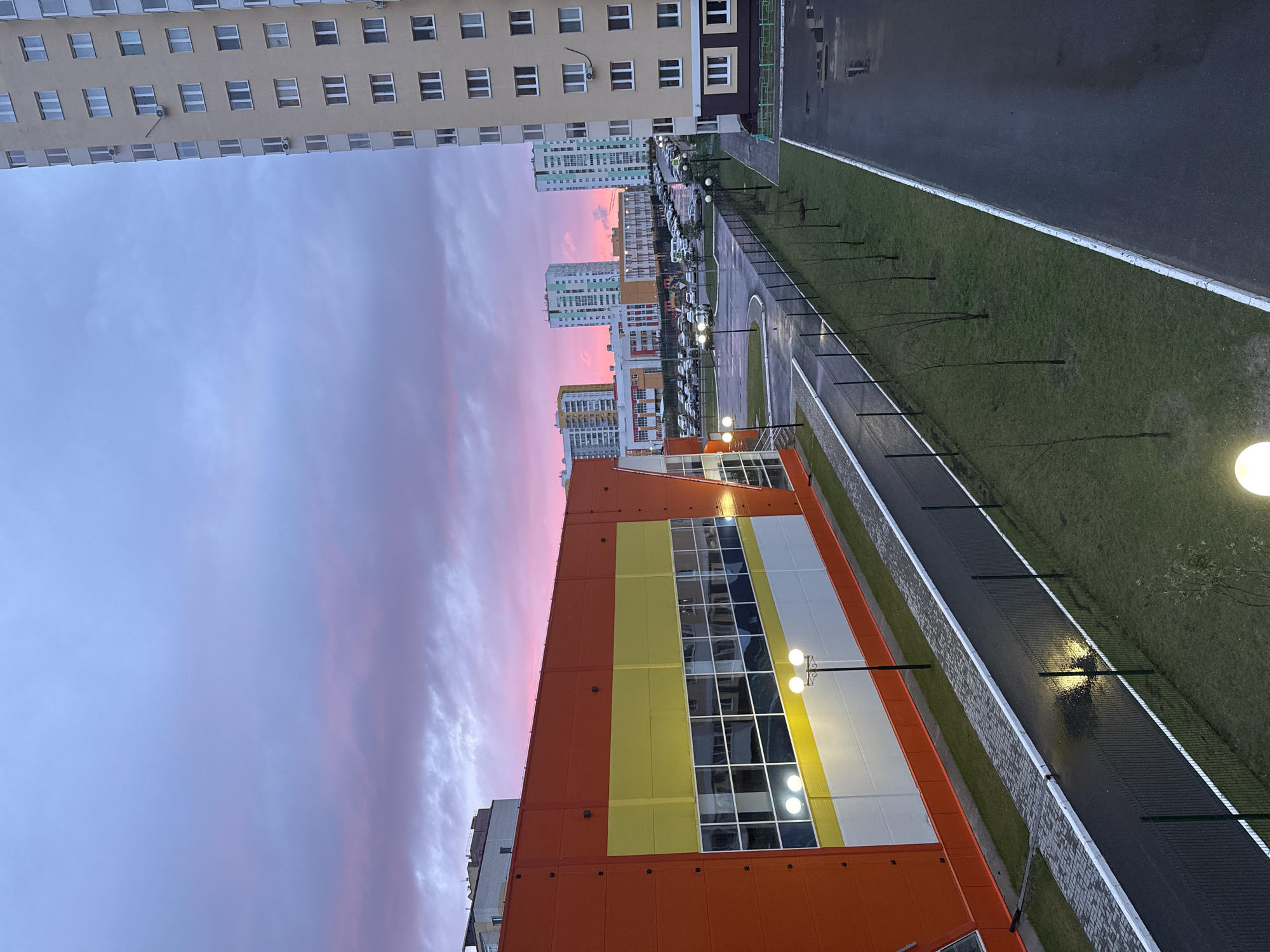 The image shows a view of Tobolsk, Russia at twilight. The sky is a blend of blue and pink hues.
Buildings dominate the skyline. A beige building with multiple windows stands on the left. Closer, a colorful building with orange, yellow, and white panels is visible. Other buildings are further away, near the horizon.
Below, there is a street with parked cars on the left. A sidewalk, a strip of grass, and a road are seen. Streetlights illuminate the area. Small trees are sparsely planted along the sidewalk. The pavement appears wet, suggesting recent rain.