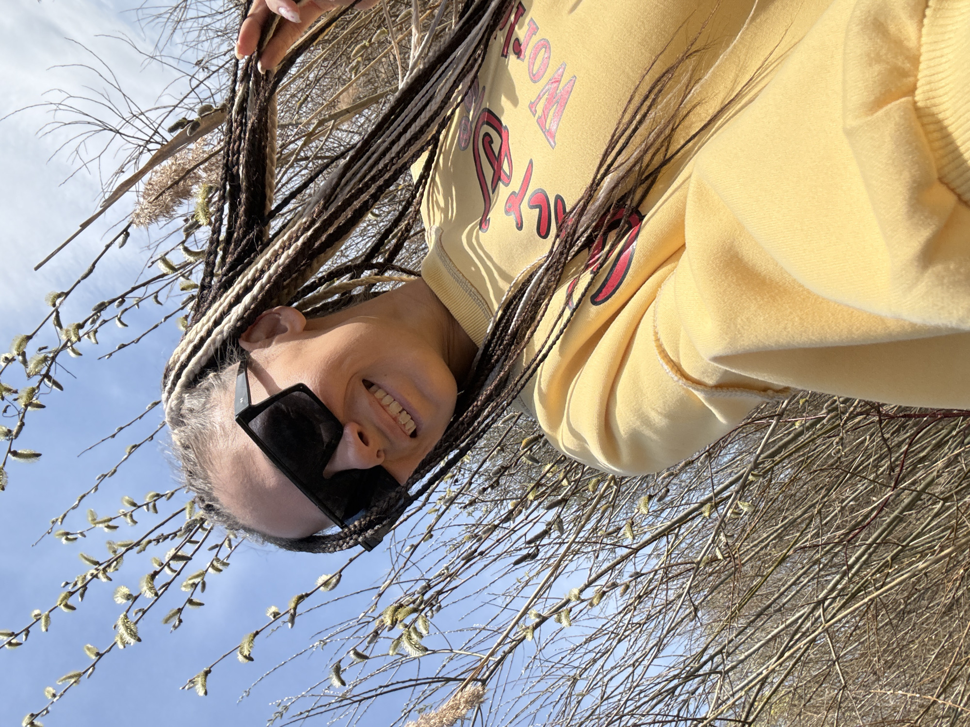 A low-angle, tilted perspective photograph shows a person with braided hair wearing black sunglasses and a yellow hoodie. The subject is smiling, facing the camera. The background consists of thin branches with small buds and a clear, blue sky. The setting is outdoors, with the person likely standing amongst trees or bushes. There is no evidence in the image to determine that the location is Rusanovo, Russia.