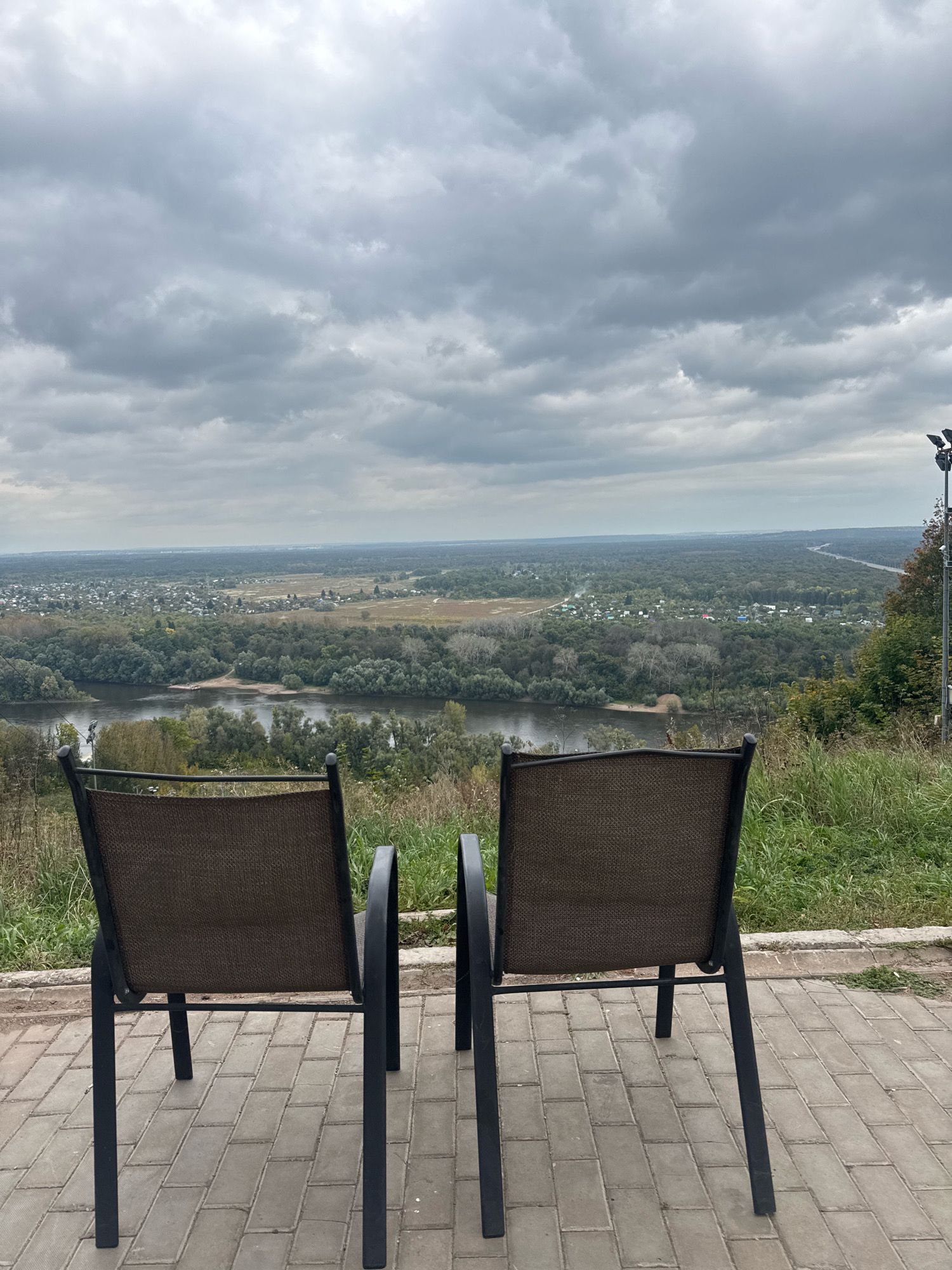The image depicts an overcast view from a high vantage point overlooking a river valley and a small town. Two empty, dark-colored chairs with metal frames and woven seats are positioned in the foreground, facing the expansive landscape. The chairs are placed on a paved walkway made of rectangular grey bricks. Beyond the chairs, the terrain slopes down towards a wide, calm river lined with lush green trees. Across the river, a settlement with numerous small houses and buildings is visible, surrounded by more dense forestation. In the distance, a highway can be seen cutting through the landscape. The sky is filled with heavy, grey clouds, suggesting a cloudy or potentially rainy day. The scene is natural and serene, with no people present. The location is identified as Ufa, Russia.