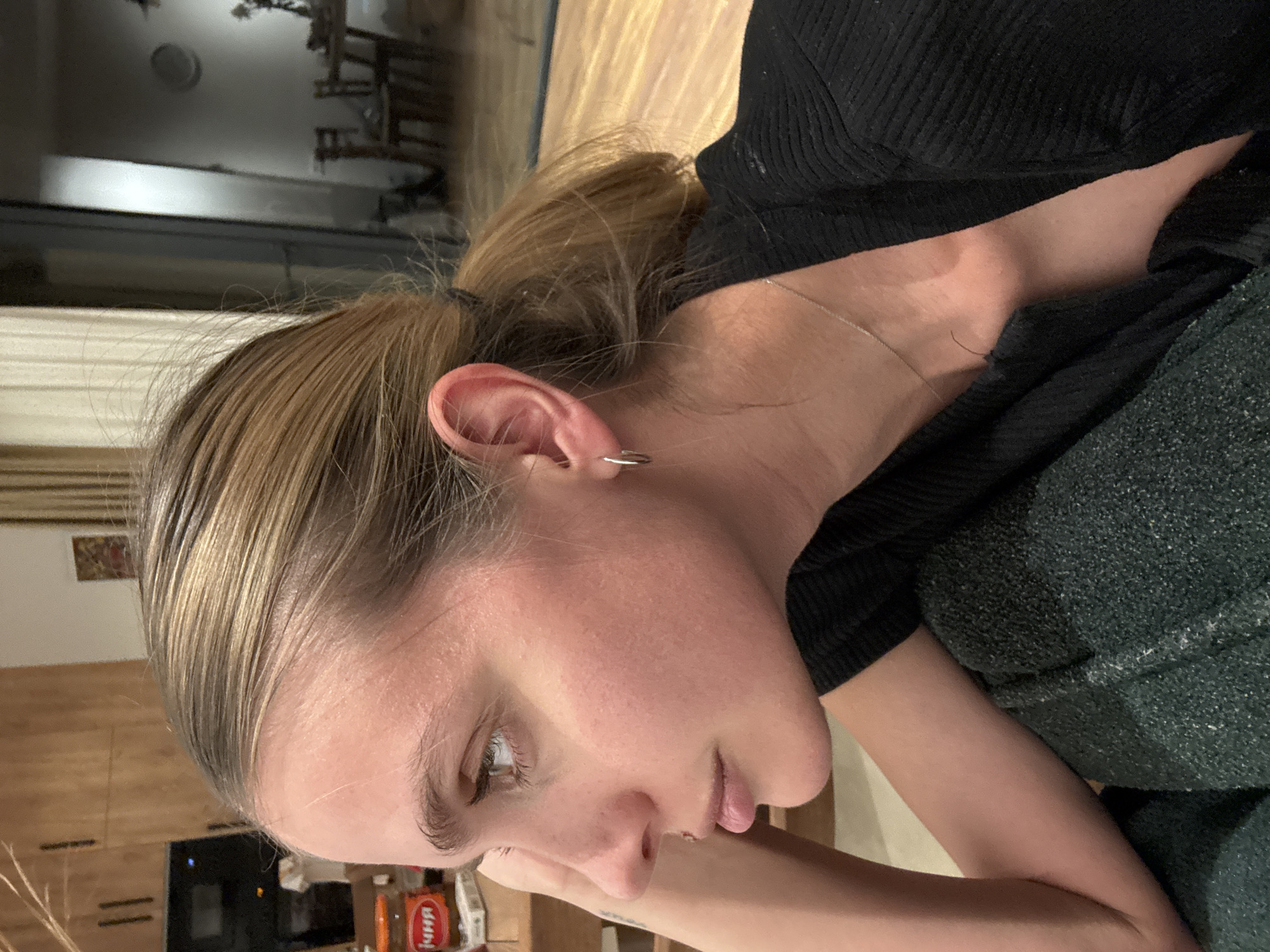 A close-up, slightly rotated image features a light-skinned female with blond hair pulled back in a ponytail. She has light brown eyes and a silver hoop earring in her left ear. She is wearing a black top and appears to be leaning on her arm. The background is somewhat blurry, but it includes a window, wooden cabinets, and a television. The location in the city of Unknown, Unknown cannot be determined from this image.
