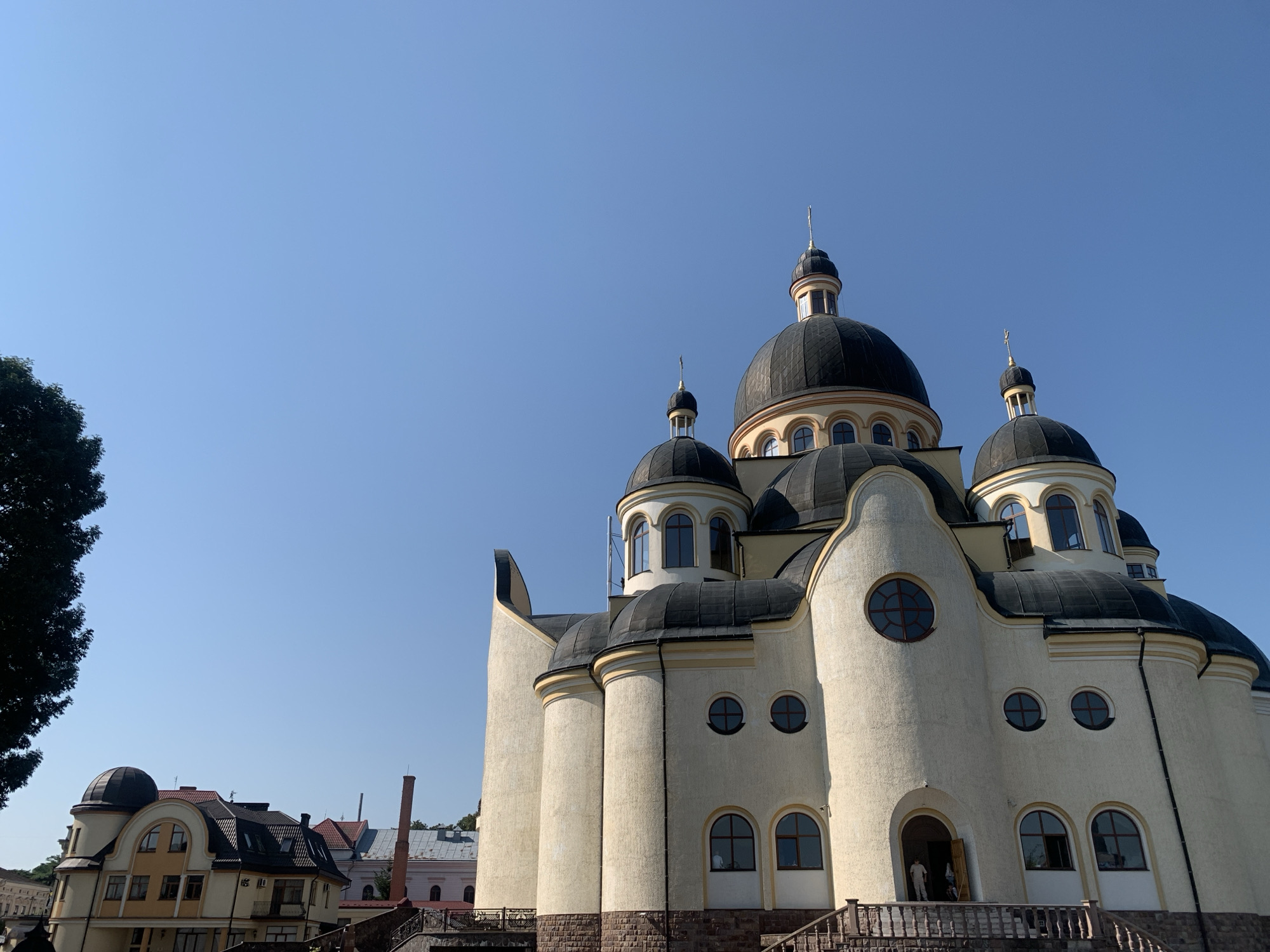 This is a low-angle, exterior, eye-level shot of the Church of the Holy Resurrection in Kolomyia, Ukraine. The large, cream-colored church features multiple domes topped with crosses, and is accented with black roofs and arched windows. The building is situated under a clear, bright blue sky. In the foreground, to the left, a dark green tree canopy is visible. In the mid-ground, to the left of the church, are several smaller buildings with red-tiled roofs. The main subject, the church, has a textured exterior with stone accents at its base. The entrance to the church is visible, with a person in a white shirt standing in the doorway. The lighting suggests it is daytime.