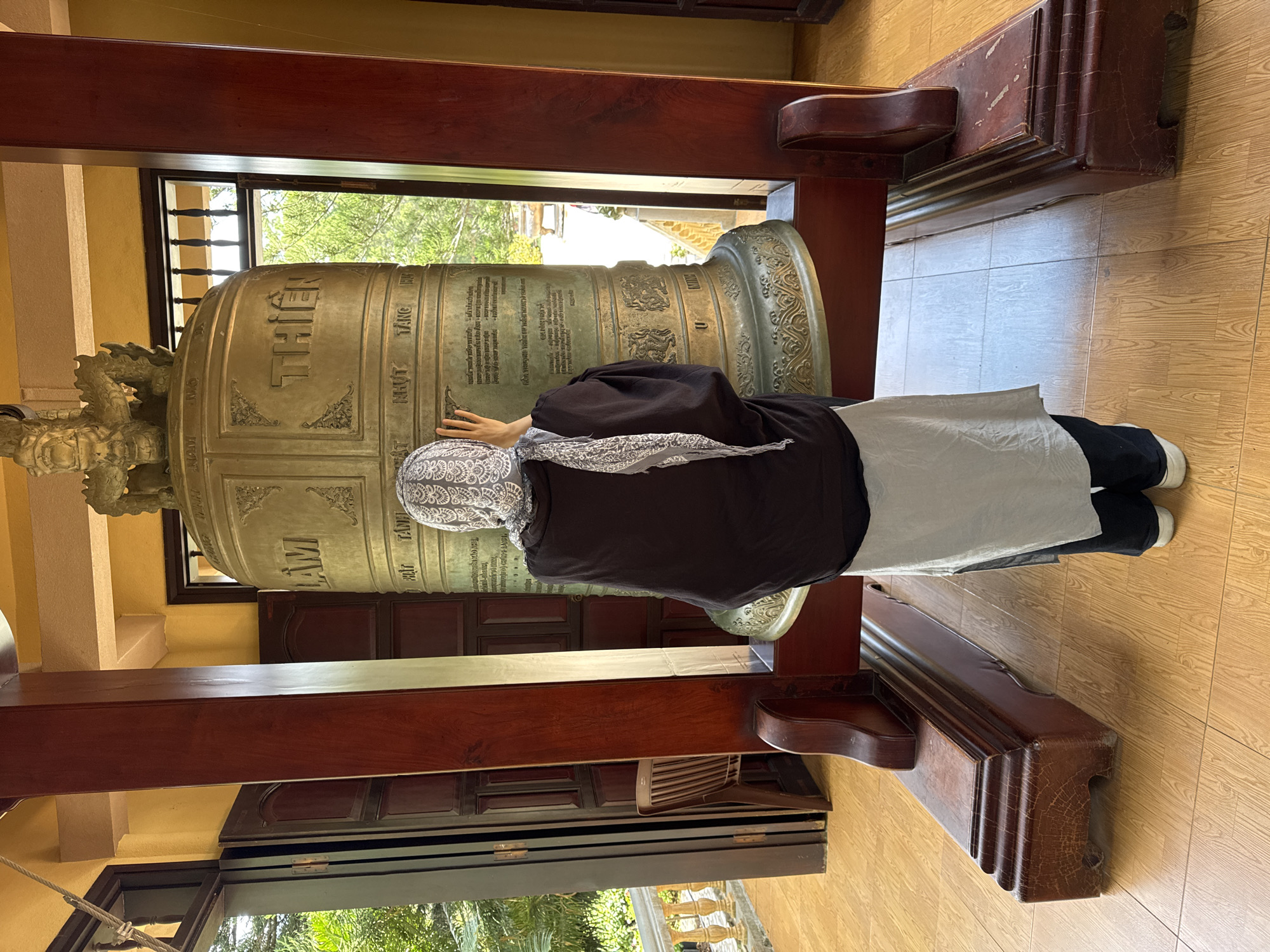 A medium shot shows a person standing in front of a large bell structure in Da Lat, Vietnam. The person, wearing a head covering with a gray and white pattern, a black top, a gray skirt, and black pants with white shoes, is touching the side of the bell. The bell is an antique bronze color with carved decorations and lettering. The bell is hung from a dark brown wooden frame with decorative carvings. The floor is a light brown wood laminate. In the background, out of a window, greenery and possibly a building can be seen.