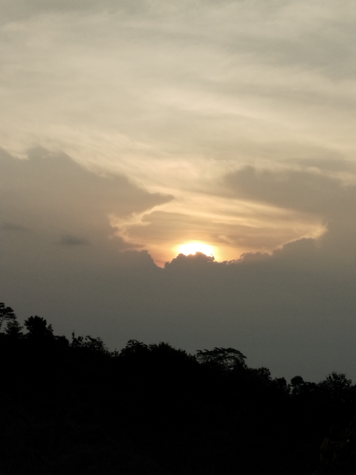 The image shows a sunset in Menjing, Indonesia. The sun is partially obscured by clouds, creating a bright halo effect. In the foreground, there is a silhouette of trees against the sky. The sky is a mix of gray and golden hues.