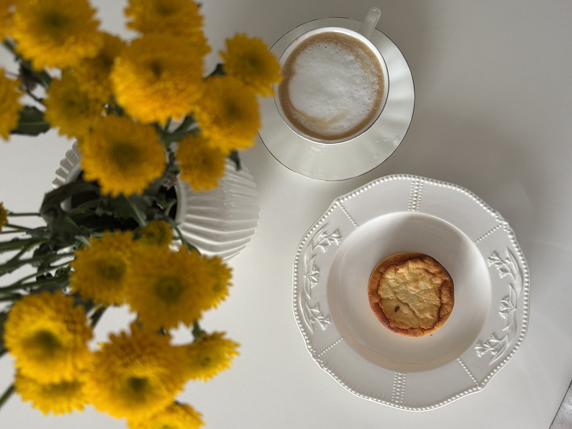 An overhead shot captures a serene still life arrangement on a bright white surface, likely a table, in what appears to be an indoor setting in Khimki, Russia. The composition features a vase of vibrant yellow chrysanthemums on the left, a cup of coffee on the upper right, and a small dessert on a plate on the lower right.
The yellow chrysanthemums, though slightly out of focus, add a splash of warm color and a natural element to the scene. They are arranged in a white, fluted or ribbed vase.
A white ceramic cup of coffee, possibly a latte or cappuccino, sits on a matching saucer with a delicate golden rim. The coffee has a thick layer of foamed milk on top, suggesting a freshly prepared warm beverage.
To the right, a single, round, golden-brown baked dessert, resembling a mini tart or cheesecake, is placed centrally on a decorative white ceramic plate. The plate features an ornate embossed pattern of dots along its scalloped edge and delicate floral designs towards the center.
The overall impression is one of calm and simple pleasure, suggesting a moment of quiet enjoyment, perhaps a coffee break or a light snack. The bright lighting and clean white backdrop create a minimalist and inviting aesthetic. There is no visible text in the image.