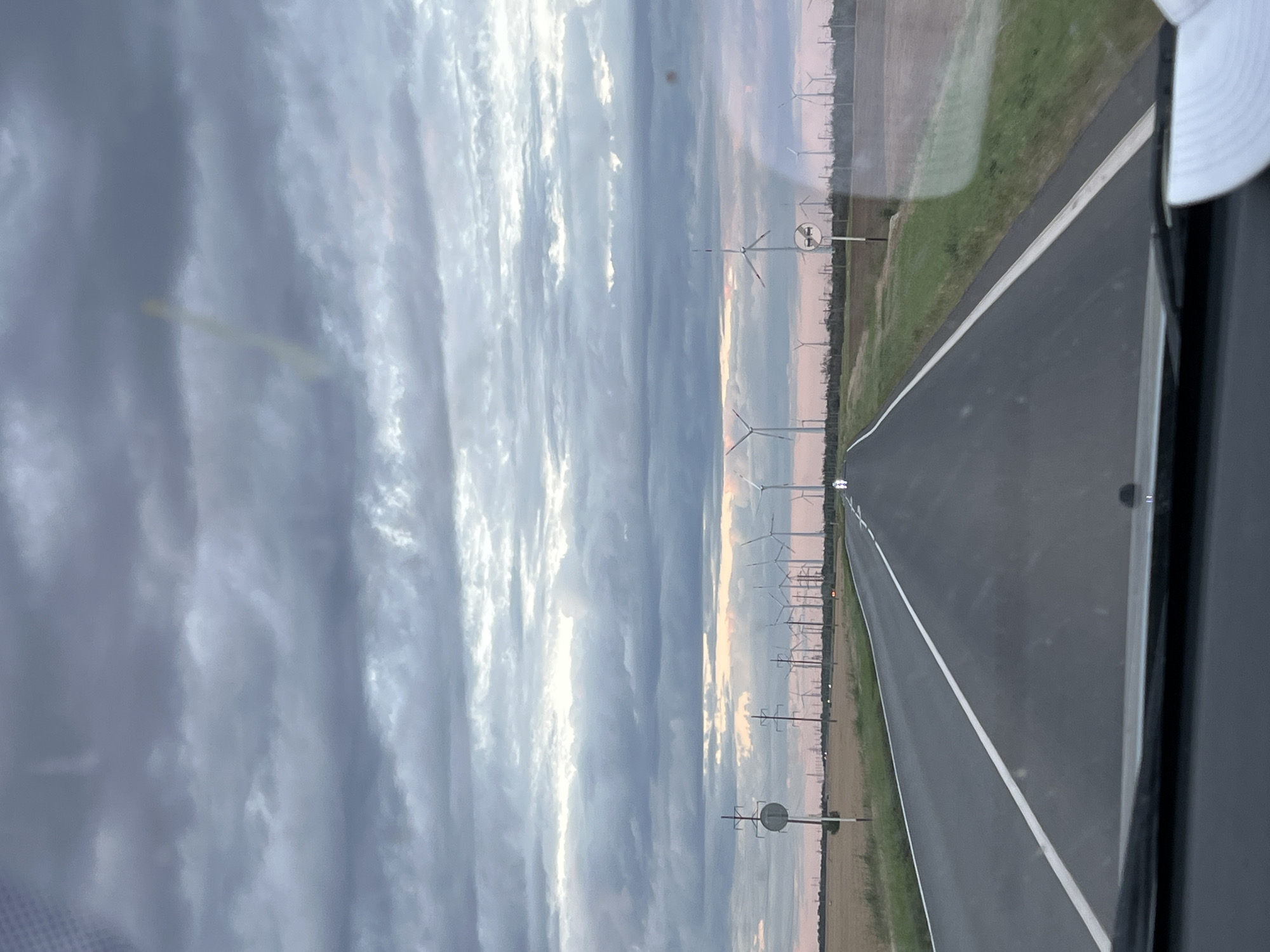 The image shows a road with a white line down the middle, bordered by a green field on the left and a line of wind turbines on the right. The sky is cloudy with patches of sunlight, suggesting it is either sunrise or sunset. The wind turbines are tall and modern, with their blades visible. There are also some power lines and poles along the side of the road. The scene appears to be rural and open, with no people or buildings immediately visible. The image is taken from inside a vehicle, as evidenced by the interior frame on the right side of the image. The overall mood is calm and serene, with a sense of vastness. The location is Zimovniki, Russia.