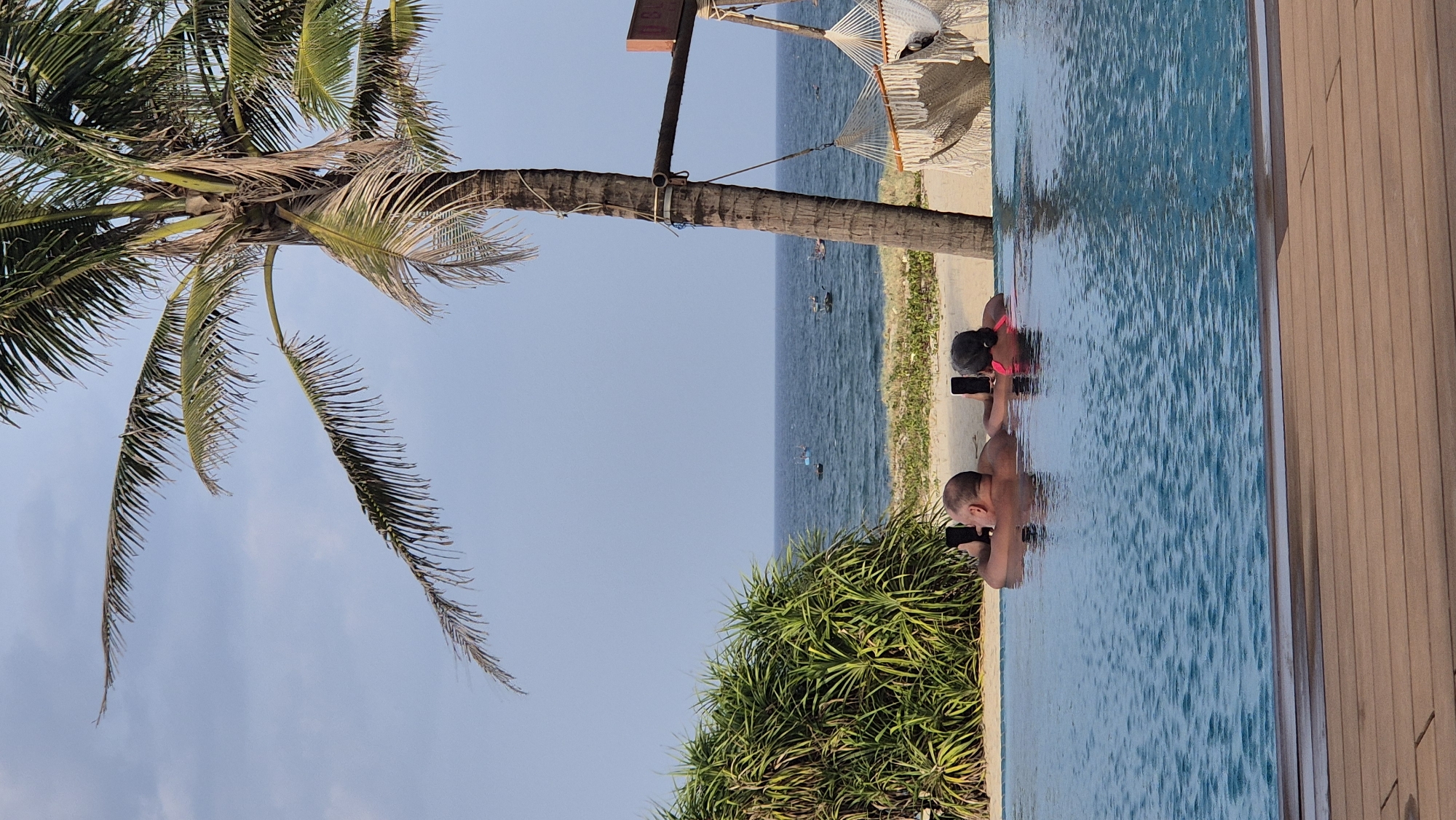 This image depicts a scene at an infinity pool overlooking the sea in Hoi An, Vietnam. Two individuals, a man and a woman, are partially submerged in the pool, each holding a smartphone. The man, facing away from the camera, appears to be bald and is wearing what looks like swimming trunks. The woman, also facing away, has dark hair and is wearing a pink top.
In the background, the sea stretches out to the horizon, with several small fishing boats visible. The coastline is lined with sand and lush green vegetation, including a prominent palm tree to the right of the frame. A hammock, suspended from a wooden structure, is also visible on the right side of the image. The sky above is a clear blue with some wispy clouds. The foreground of the image shows the edge of the infinity pool and wooden decking. The overall impression is one of a relaxed tropical resort setting.