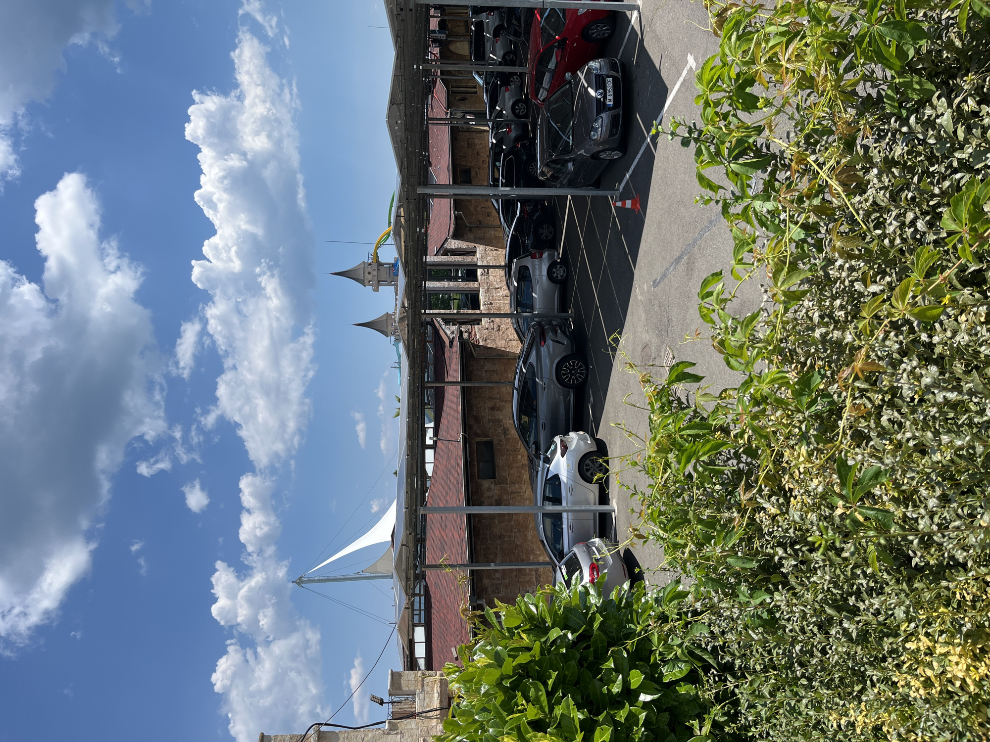 The image depicts a parking lot with several cars parked under a covered structure. The structure has a metal frame with a translucent roof, possibly designed to provide shade or shelter. The parking lot is paved, and white lines mark the parking spaces. In the background, there are buildings with reddish-brown roofs, and architectural elements resembling towers or spires, suggesting a resort or themed area. A large white sail-like structure is visible in the distance. Lush green vegetation, including bushes and climbing vines, frames the foreground of the image, obscuring some of the parked cars. The sky is blue with scattered white clouds. The scene appears to be a sunny day in Ravda, Bulgaria.
