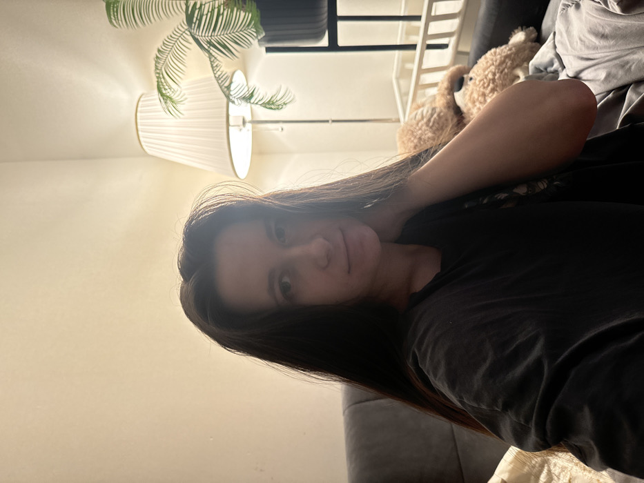A person with long, dark hair rests on a gray couch. A floor lamp with a pleated shade illuminates the scene, positioned near a plant. In the background, a teddy bear sits on a white shelf. The person is wearing a dark-colored top. The lighting is soft, creating a cozy atmosphere.