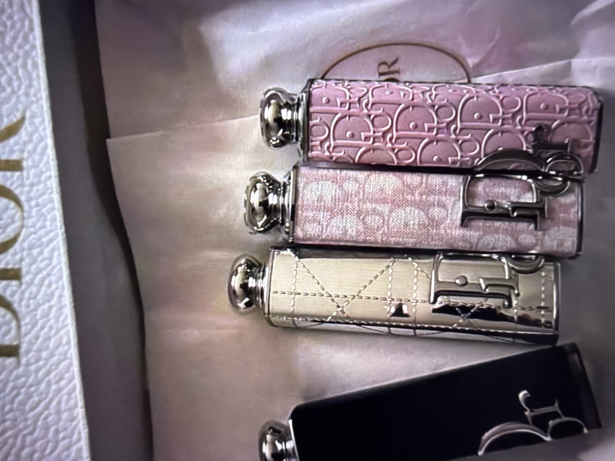 The image shows a close-up, overhead view of four Dior lipstick cases arranged in a box. Three of the lipsticks are light-colored with ornate silver caps and clasps featuring the Dior logo. The top two lipsticks have a repeating Dior pattern, one in a pale pink and the other in a muted pinkish-white. The third lipstick is a pale ivory color with a quilted design. The fourth lipstick, at the bottom of the frame, is black with silver accents. The box is white with a textured pattern and embossed gold lettering that appears to say "DIOR" on the left side. The overall presentation suggests a new or curated collection of luxury cosmetics. The lighting is soft and even, indicating an indoor setting, likely a retail or personal display. No people are visible, and no specific activity is taking place beyond the arrangement of the objects. The image focuses solely on the products themselves and their packaging. The location is Beșalma, Moldova, which provides a geographical context but no visual cues within the image itself.