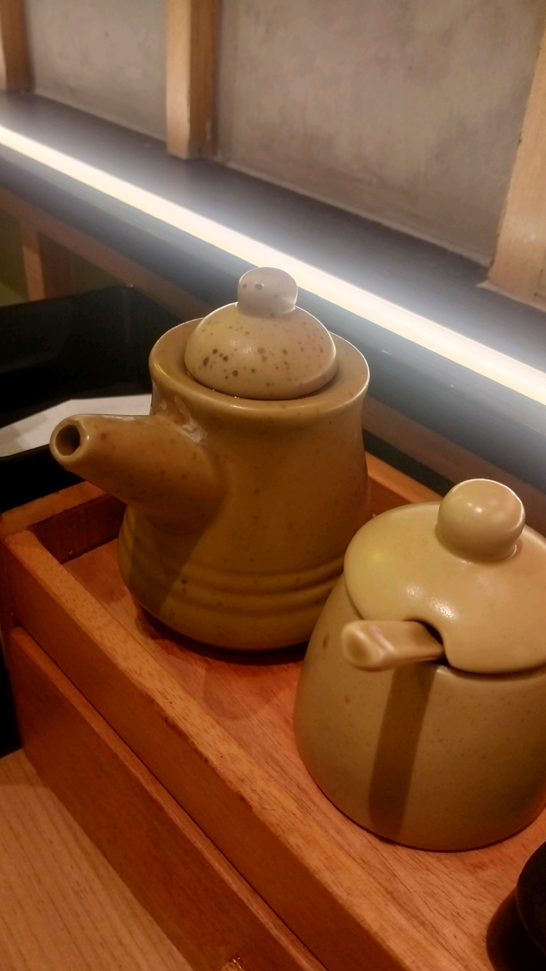 Two ceramic condiment containers are placed on a wooden tray. The container on the left is a small teapot with a spout and a lid. The container on the right is a lidded jar with a small spoon protruding from its lid. The containers are a light tan color and appear to have a speckled finish. They are situated on a wooden surface that is part of a tray with raised edges. The background shows a wall with wooden beams and a strip of illumination. The scene is indoors, likely in a restaurant or dining area. There are no people visible in the image. The image does not provide any information that would indicate its location in Lhokseumawe, Indonesia.
