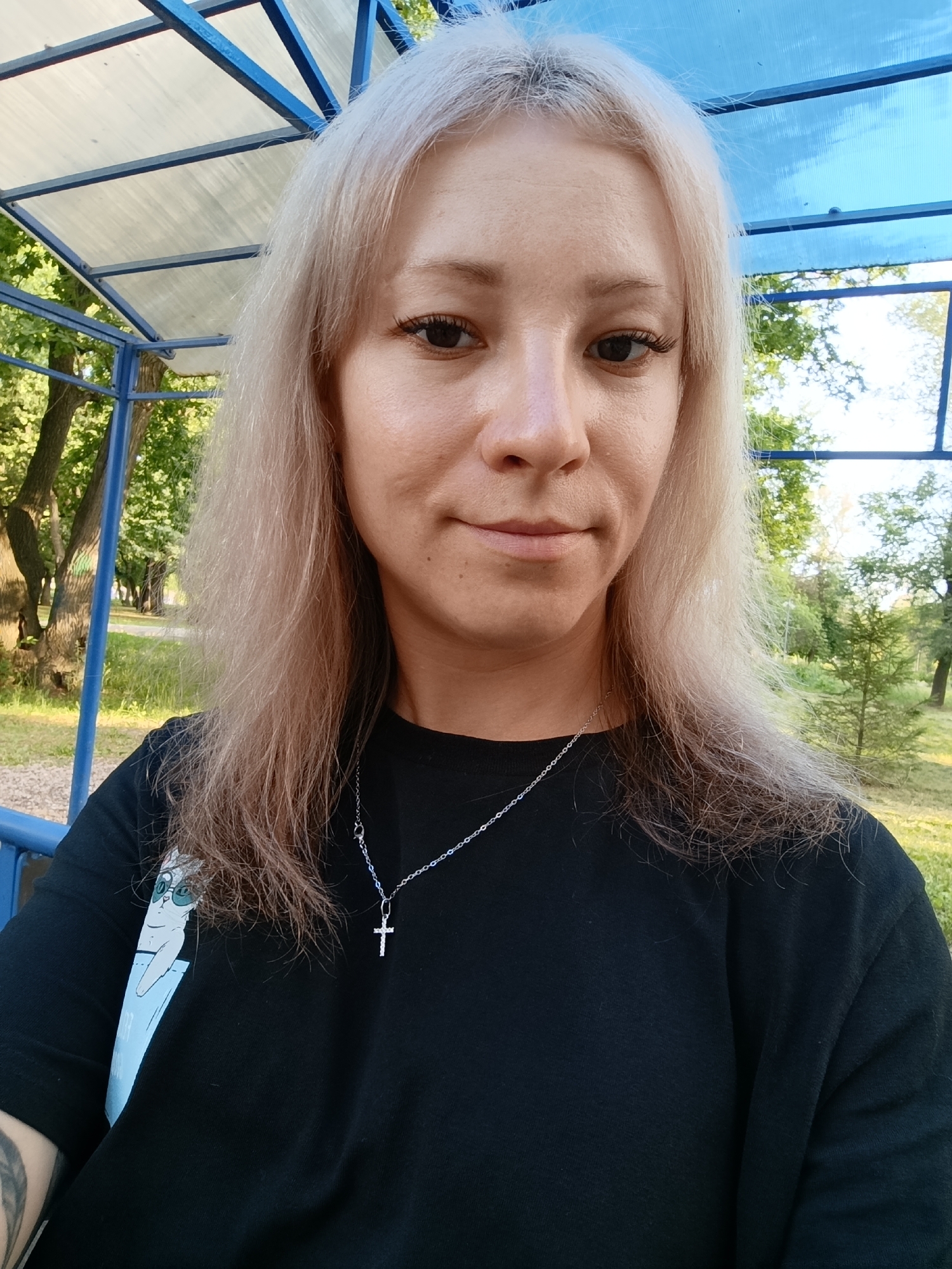 A blonde woman with medium-length hair, wearing a black t-shirt and a silver cross necklace, is pictured sitting outside under a blue and clear covered structure. She is looking at the camera with a slight smile. The background shows a park with trees and green grass. The structure appears to be a bus stop or a pavilion in a public park.
