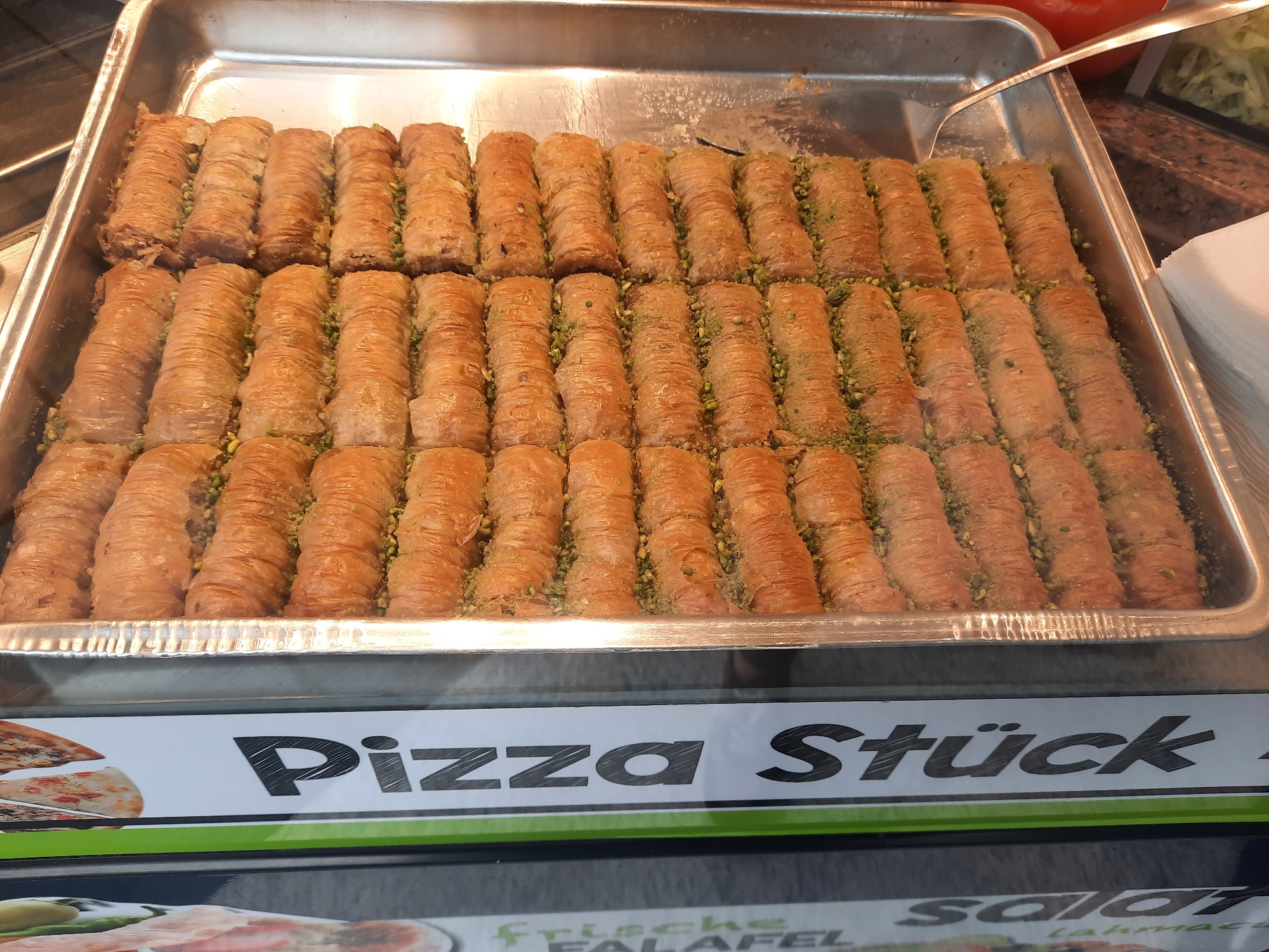 The image shows a close-up, eye-level view of a metal tray filled with rows of baklava in a food establishment. The baklava are golden brown, cylindrical, and appear to be made with layers of phyllo dough and a filling, likely nuts, with visible green pistachio fragments between the layers. They are arranged in neat rows. A metal serving utensil is partially visible resting on the edge of the tray. Below the tray, a white sign with black lettering displays "Pizza Stück" with a graphic of pizza slices on the left. The background includes blurred elements of a food display and a glass counter. The scene is likely a bakery, pastry shop, or deli counter, possibly in Rosenheim, Germany, given the German text on the sign.