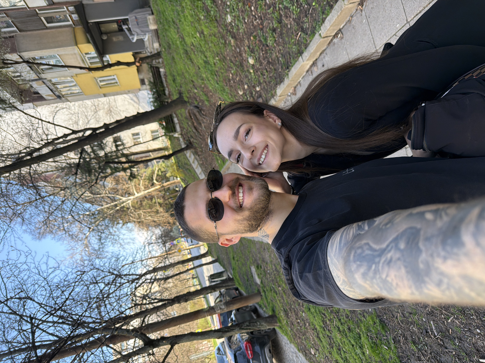 A selfie shows a smiling couple standing on a sidewalk in Sofia, Bulgaria. The man is holding the camera and is wearing sunglasses and a black t-shirt, his left arm is heavily tattooed. The woman is next to him wearing sunglasses on her head and a black blouse, smiling. They are in front of a grassy area with bare trees and some parked cars on the street. Buildings can be seen in the background with the sky visible above the trees. The picture is rotated slightly counter-clockwise.