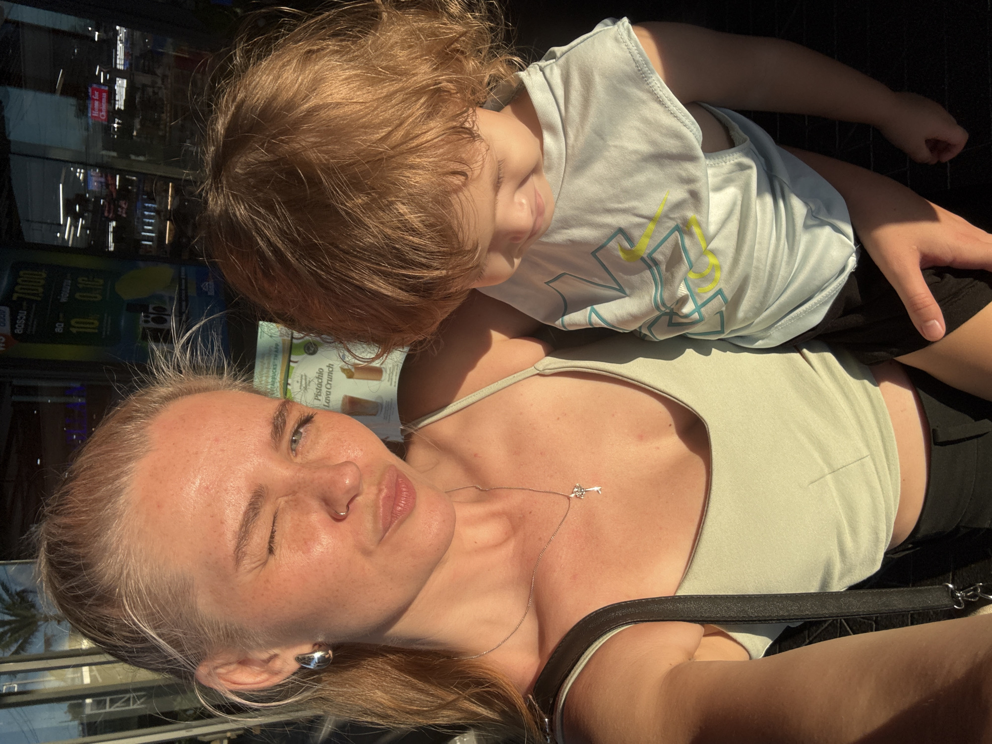 A young woman with blonde hair pulled back in a ponytail takes a selfie outdoors. She is wearing a light green tank top and black pants. She has a silver earring in her left ear and a necklace with a key pendant. She is winking and puckering her lips. She is holding a young child with reddish-brown curly hair in her arms. The child is wearing a light blue t-shirt with a yellow and teal graphic. In the background, there is a building with a glass facade, displaying some signs and posters. One sign appears to be for a coffee shop, mentioning "Pistachio Lava Crunch." The scene is bathed in warm, late afternoon sunlight, casting long shadows. The overall impression is a candid moment captured during an outing. The scene is consistent with an urban or commercial area, potentially a shopping or dining district. The presence of palm trees reflected in the glass suggests a tropical location. The currency displayed on one of the signs (Baht) confirms the location as Thailand. The specific location within Choeng Thale, Thailand is not identifiable from the visual information.