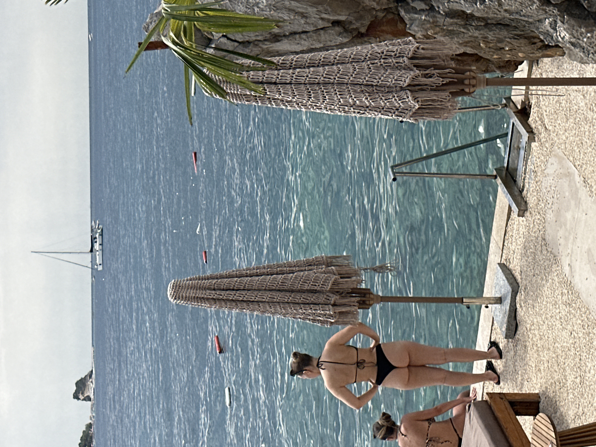 The image depicts a seaside scene in Budva, Montenegro, with two women in bikinis standing near the water. The woman in the foreground, facing away from the camera, is wearing a black bikini and has her hands on her hips. To her left, another woman in a patterned bikini is seated. Two large, tasseled umbrellas stand on the paved area. The turquoise water of the Adriatic Sea stretches out, with a sailboat visible in the distance and a rocky outcrop to the right. The setting appears to be a beach or waterfront promenade, characterized by a pebbled ground and a metal railing leading into the water.