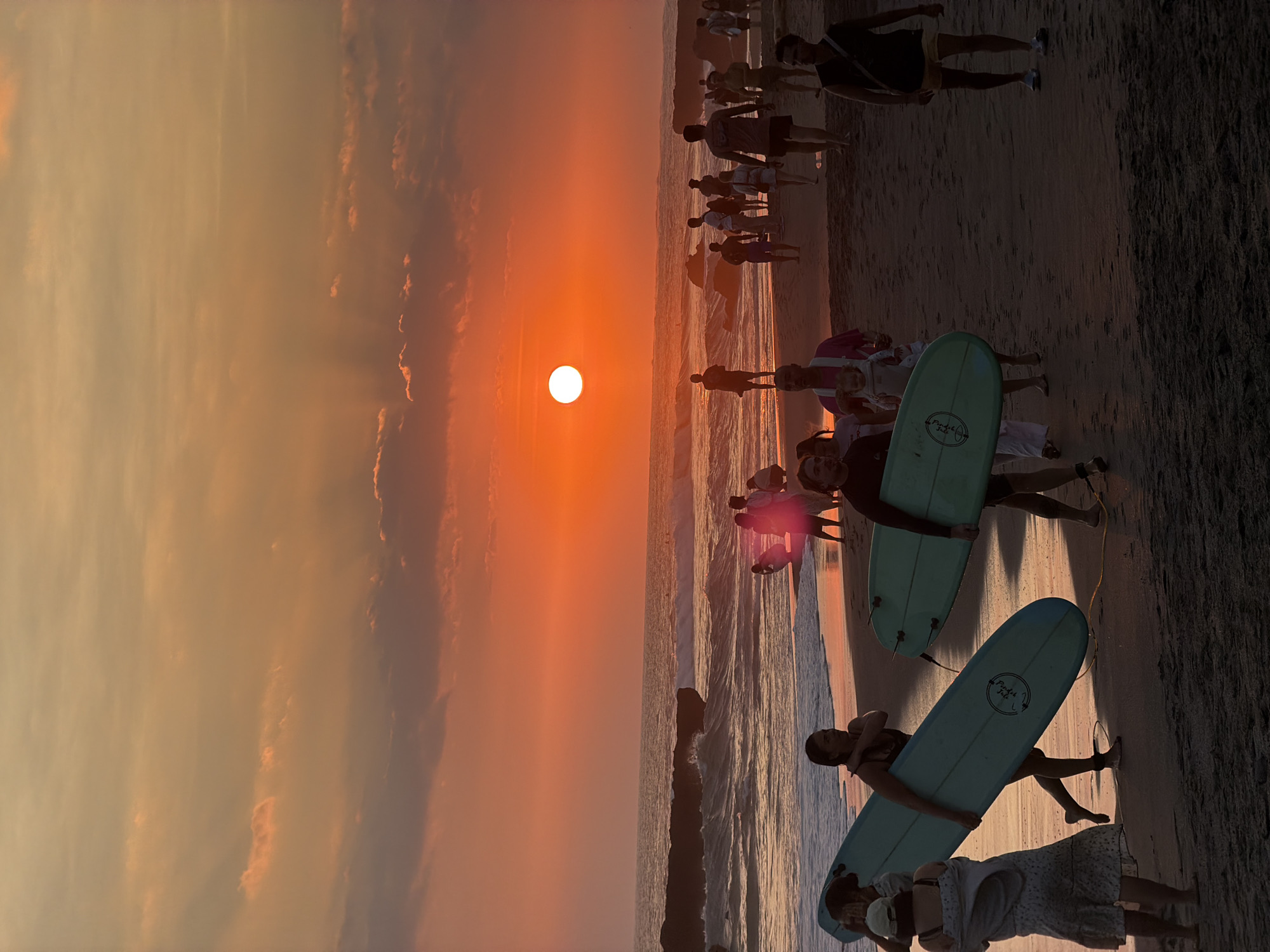 A group of people walk on a beach at sunset in North Kuta, Indonesia. The sun is setting over the ocean, casting a warm orange glow over the scene. Several people are carrying surfboards, suggesting they have been surfing or are about to go surfing. The waves are visible in the background, with some rocky formations breaking up the surf. The sand on the beach is dark, and the sky is filled with dramatic clouds. Some of the people are silhouetted against the setting sun, while others are more clearly visible, walking and interacting with each other. The overall atmosphere is one of leisure and enjoyment.