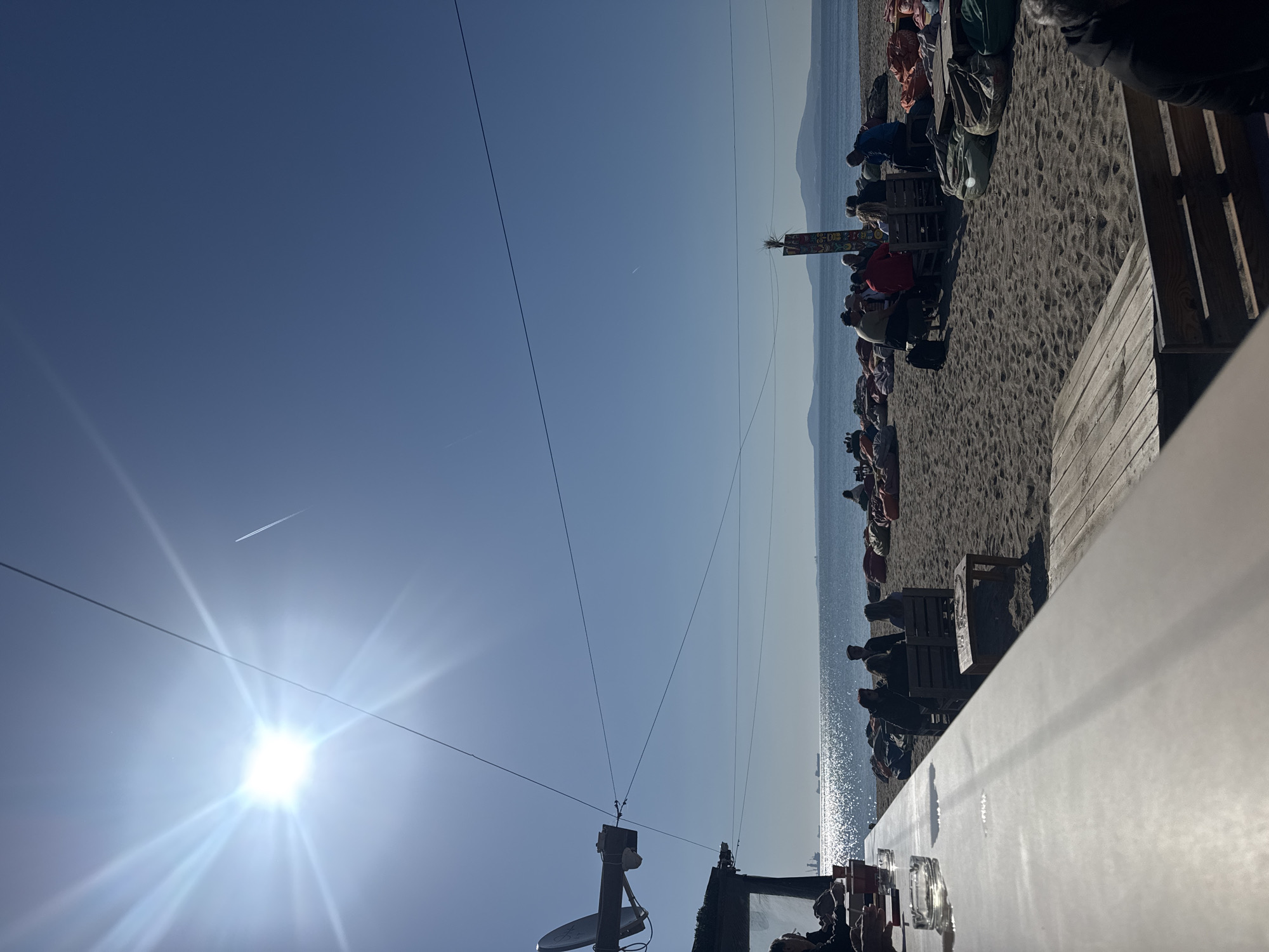 The image depicts an outdoor scene at a beach in Burgas, Bulgaria, on a sunny day. The foreground shows a partially visible table with some objects on it, possibly located in a restaurant or cafe setting. People are seated at tables on the beach, likely customers. Behind them, the sea stretches to the horizon, with mountains visible in the distance. The sky is clear and blue, with the sun shining brightly. Power lines run above the scene. An airplane contrail is visible in the sky.