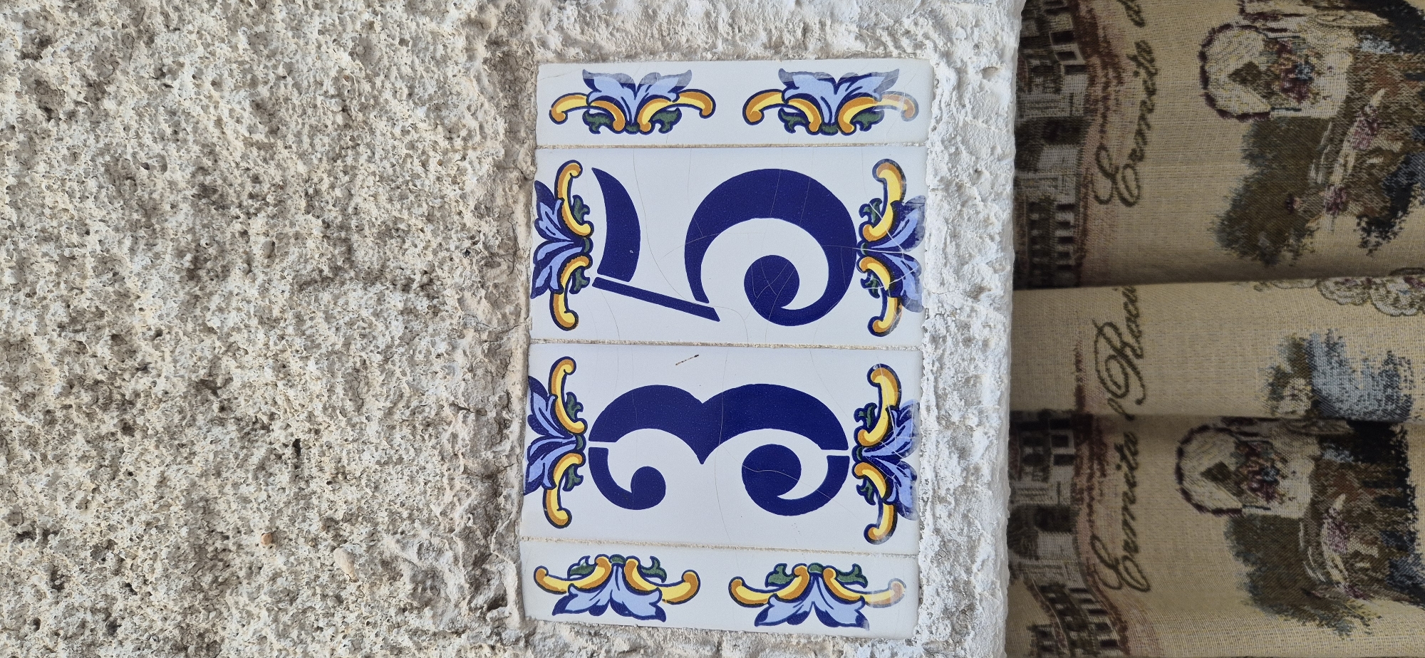 A close-up, eye-level shot shows a house number "35" displayed on two ceramic tiles, set against a textured, light-colored stone wall. The tiles are white with blue numbers and decorative yellow and blue floral patterns on the edges. The "3" is on the left tile and the "5" is on the right tile. The number "35" is framed by decorative blue and yellow floral motifs. Below the tiles, a tapestry or curtain with a landscape and text, possibly "Ermita del Rocio", is visible. The lighting is neutral, suggesting daylight. The scene is static and outdoors.