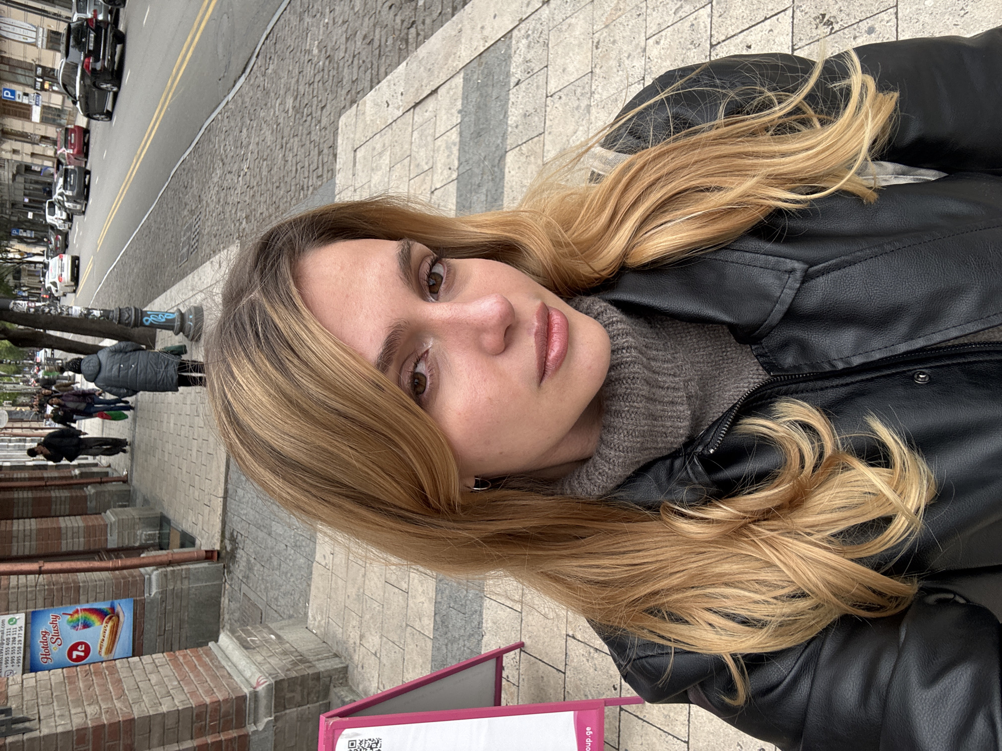 A young woman with long, wavy blonde hair and brown eyes is featured prominently in what appears to be a selfie taken on an urban street in Tbilisi, Georgia. She looks directly at the camera with a neutral expression, wearing a black leather jacket over a grey ribbed knitted sweater. Her hair cascades over her shoulder and arm.
The background reveals a bustling city scene. A wide sidewalk, composed of stone tiles and cobblestones, stretches behind her. To the left, a street is visible with parked cars and more vehicles in the distance, indicating traffic. Pedestrians are seen walking further down the sidewalk, suggesting a normal daytime street activity. Buildings with facades of brick and light-colored stone line the street. A black lamppost with some blue graffiti is also visible.
On the right side of the image, a vertical billboard or stand displays text including "Hotdog Slushy" with a rainbow graphic and "7a". Below this, several phone numbers beginning with the Georgian country code "+995" are listed: "+995 555 406 111", "+995 555 280 111", and "+995 558 29 77 56". An incomplete email address ending "@gmail.com" and a partially visible website "oup.ge" (with '.ge' being Georgia's country code top-level domain) are also present, along with a QR code. A "P" parking sign is also visible in the background. The lighting is soft and diffused, suggesting it's daytime, possibly under an overcast sky.