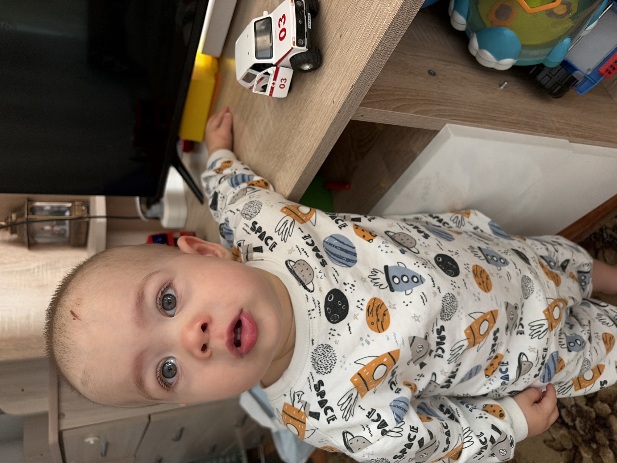 A baby wearing a white pajama set with a space-themed design, featuring planets, rockets, and the word "space", is lying on a carpet near a shelf with toys. The child has grey eyes, fair skin, and a small scrape on his forehead. On the shelf, next to the baby's hand, is a white toy car labeled "03" and a blue and yellow toy. A portion of a television is visible in the background. The room has light brown wooden surfaces. This photo was likely taken in a residence in Chaikovsky, Russia, based on the "LADA 4x4" branding on the toy car.
