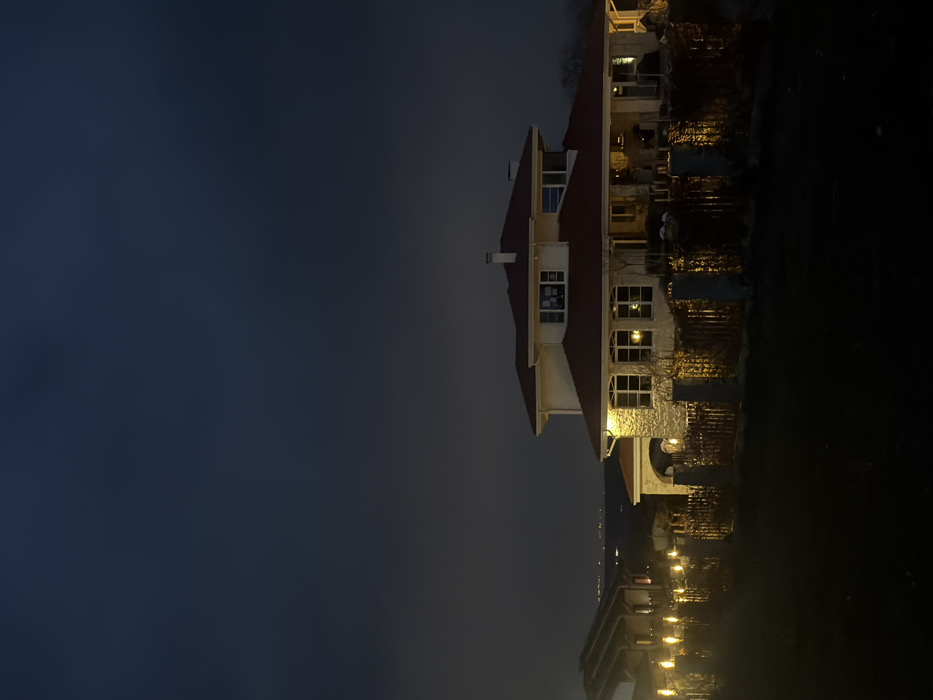 This image depicts a nighttime scene in Priseltsi, Bulgaria, featuring a large, two-story residential house and its surroundings under a dark, overcast sky. The prominent house, situated slightly right of center, exhibits a light-colored (possibly stone or stucco) facade on its lower level and a lighter upper story, topped with a distinct red tiled roof and a visible chimney. Multiple windows are present, with several on both levels illuminated from within, indicating interior lights are active. The front of the house features an illuminated porch or covered patio area.
In the mid-ground, a wooden fence with regularly spaced light-colored posts stretches across the frame, positioned in front of the main residence and extending to the left. Behind this fence, dark hedges or shrubs are visible, reflecting ambient light. A string of bright, vertical, torch-like or flame-effect lights are prominently displayed along the left segment of the fence line. To the far left, a portion of another structure, possibly another house or auxiliary building with a similar roof, is partially visible. The immediate foreground consists of dark, unidentifiable ground cover. In the distant background, faint city lights are visible along the horizon, suggesting an elevated vantage point. No individuals or specific actions are discernible within the frame.