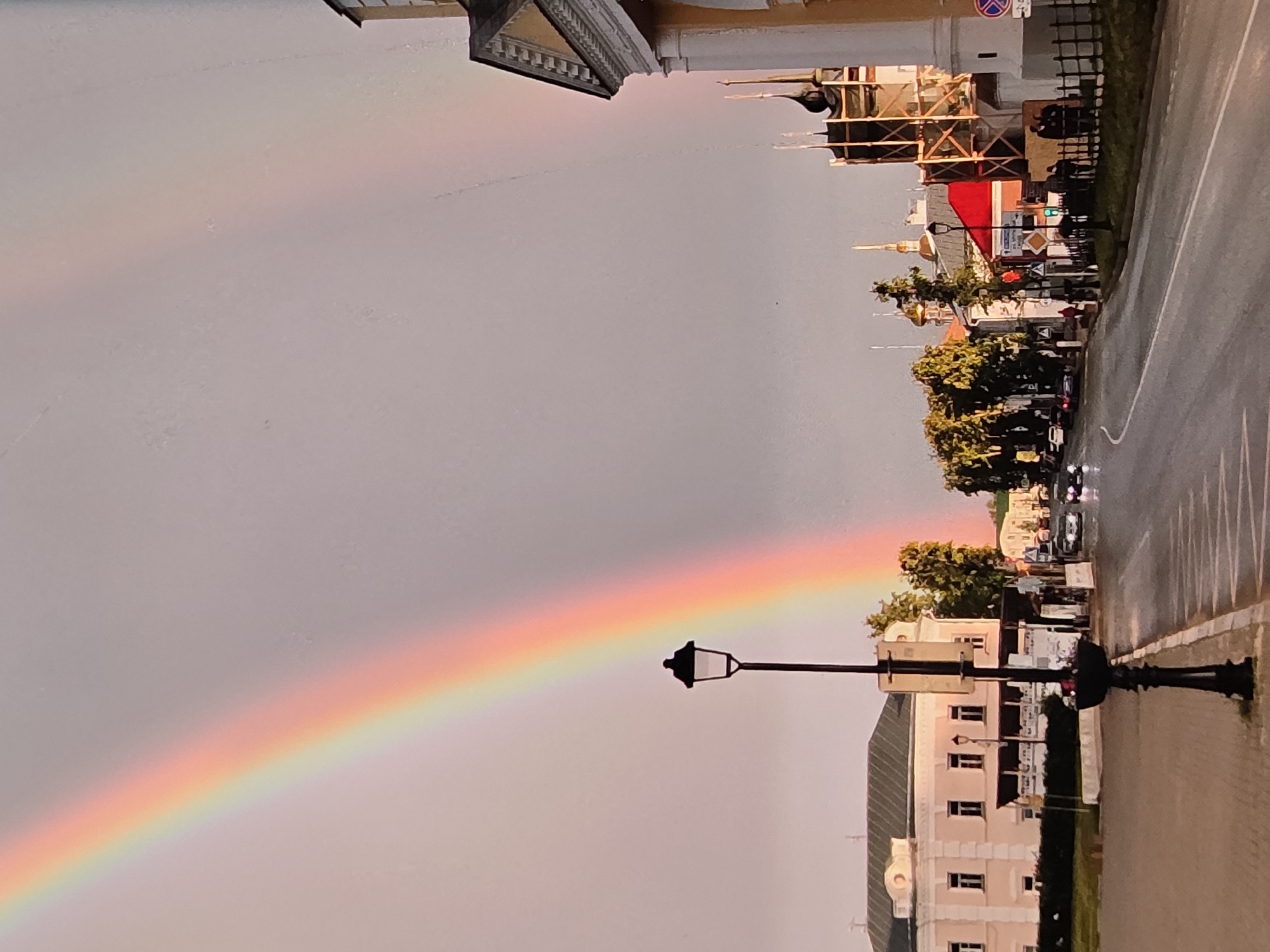 The image depicts a street scene in the city of Vladimir, Russia, with a prominent rainbow arcing across a pale sky. The rainbow is visible on the left side of the frame, with its colors fading into the light sky. In the foreground, a black lamppost with a decorative base stands on the right side of the street, with a flower planter at its base. The street is paved and wet, suggesting recent rain.
To the left of the street, a light-colored, multi-story building with a dark roof and numerous windows is visible. A row of trees with green foliage lines the edge of the street in front of this building. Further down the street, other buildings can be seen, including one with a red roof and another with architectural features characteristic of a church, including domes and spires. Some of these buildings appear to be under construction, with scaffolding visible around one.
Several cars are present on the street, some in motion. Pedestrians can be seen on the sidewalks and near the buildings, including a group of people standing near the fenced area of the church-like building. The overall atmosphere of the scene is calm, with the natural phenomenon of the rainbow adding a touch of wonder to the urban environment.