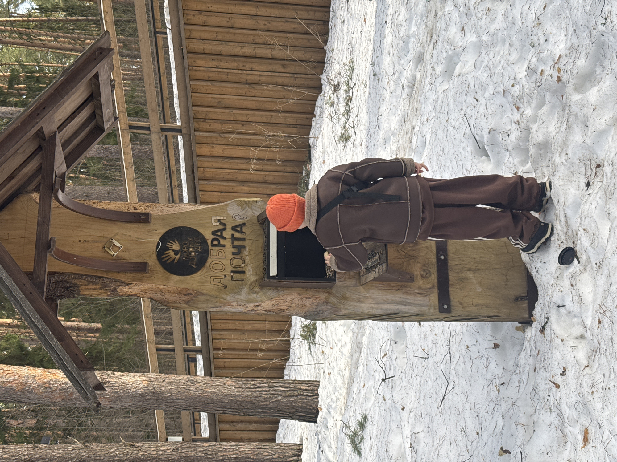 A child wearing a brown jacket, brown pants, an orange hat, and carrying a black shoulder bag, peers into a mailbox labeled "ДОБРАЯ ПОЧТА?" (Good Mail?) in Votkinsk, Russia. The mailbox is constructed of wood, possibly from a tree trunk, and has a sloped roof. The ground is covered with snow, and a wooden fence is visible in the background, adjacent to tall trees.