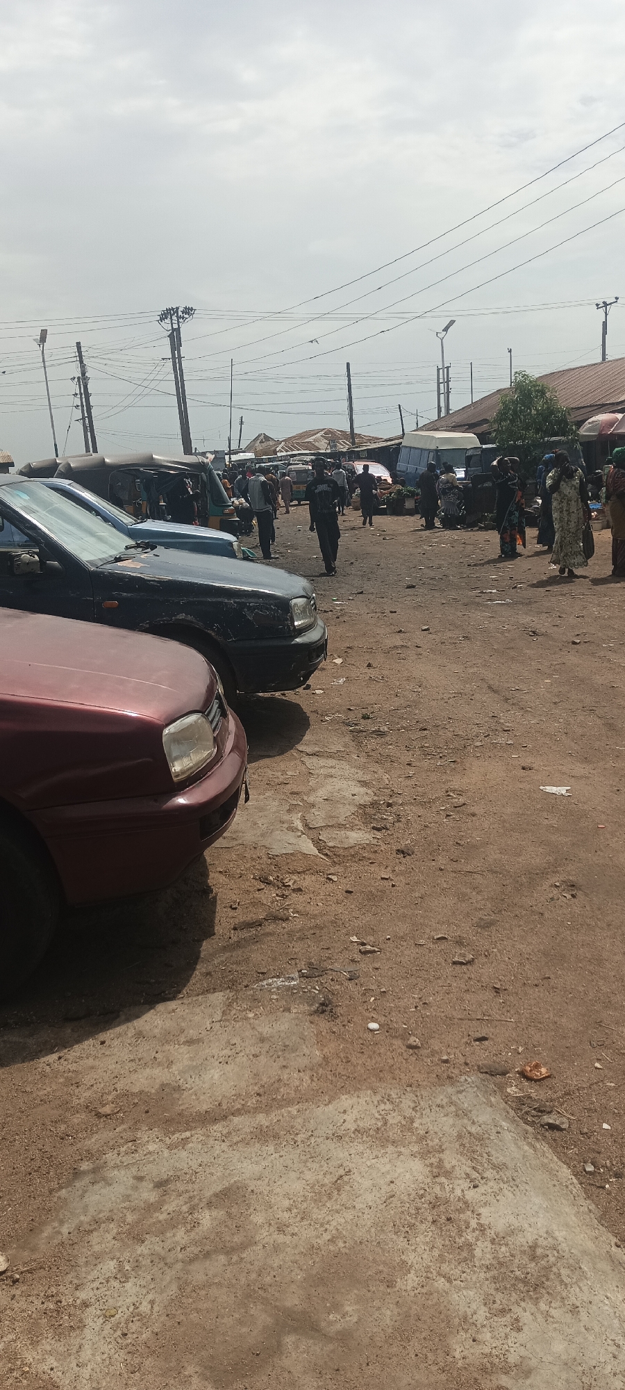 The image depicts a bustling scene at a market in Bukuru, Nigeria. In the foreground, three cars of varying colors (maroon, dark blue, and light blue) are parked on a dirt lot with areas of exposed concrete. The ground is uneven, littered with small debris. Further back, a crowd of people is visible, some walking and others standing, suggesting active market stalls. Several vehicles are interspersed within the crowd, including a yellow motorized tricycle (keke), a white van, and cars. In the background, corrugated metal roofs cover market stalls or buildings. Utility poles with power lines crisscross the sky. The overall scene suggests a busy, somewhat congested market environment under a cloudy sky.