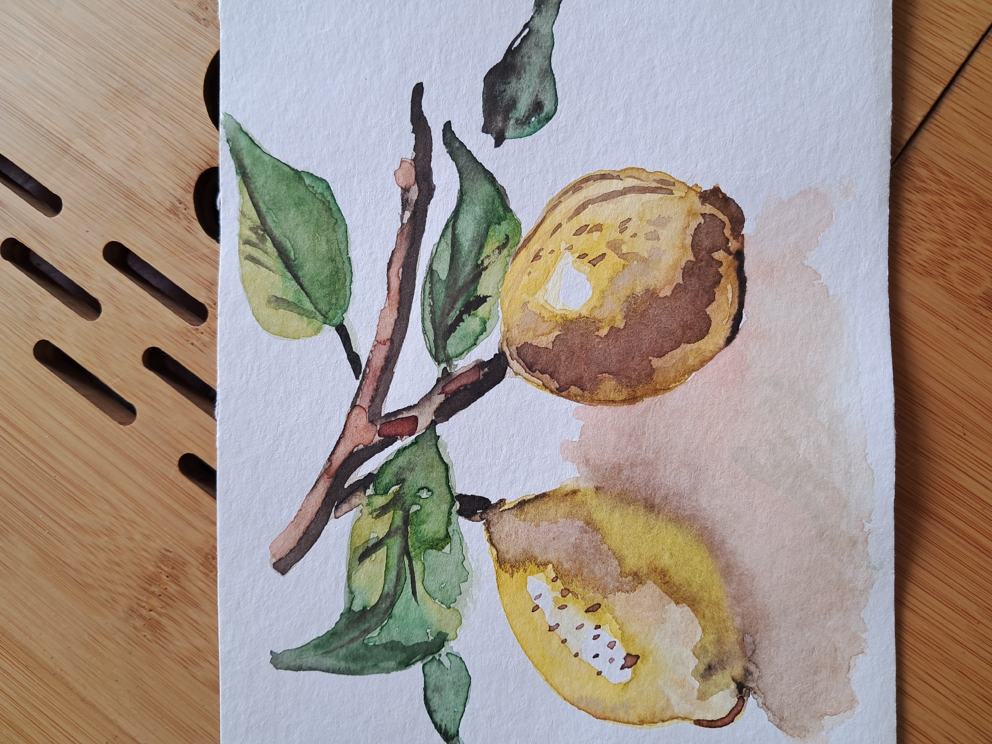 A watercolor painting depicts two lemons on a branch with leaves against a white background. The lemons are rendered in shades of yellow and brown, with visible brushstrokes and watercolor effects like bleeding and pooling. One lemon is cut in half, revealing white pulp and brown seeds. The leaves are painted in varying shades of green, with dark brown veins. The branch is brown and textured. A soft, pale pink and brown wash creates a subtle shadow beneath the lemons. The painting is placed on a light brown wooden surface with parallel grooved lines.