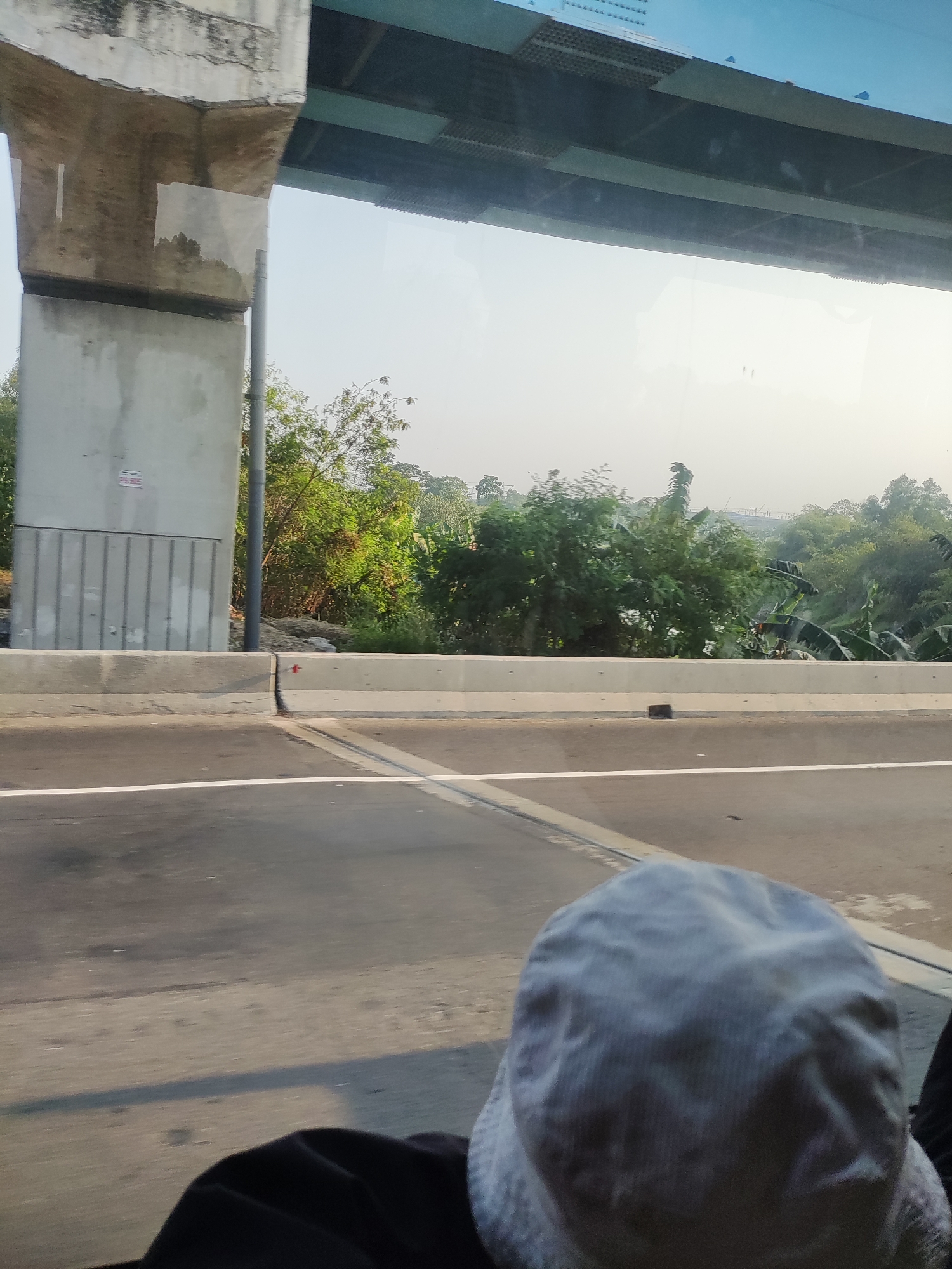 This image captures a view from a vehicle, likely a bus or car, traveling on a road near Karawang, Indonesia. In the foreground, the back of a person's head wearing a light-colored hat is visible. The road surface, marked with a white line, is textured and shows signs of wear. To the left, a large concrete pillar supports an elevated structure, possibly a bridge or an overpass, which extends into the background. This structure has a darker colored underside with visible grating. Beyond the road and the pillar, a lush green landscape with various trees and foliage is present. The sky is hazy and appears to be a bright, overcast day. The scene suggests an urban or semi-urban environment with ongoing construction or infrastructure development.