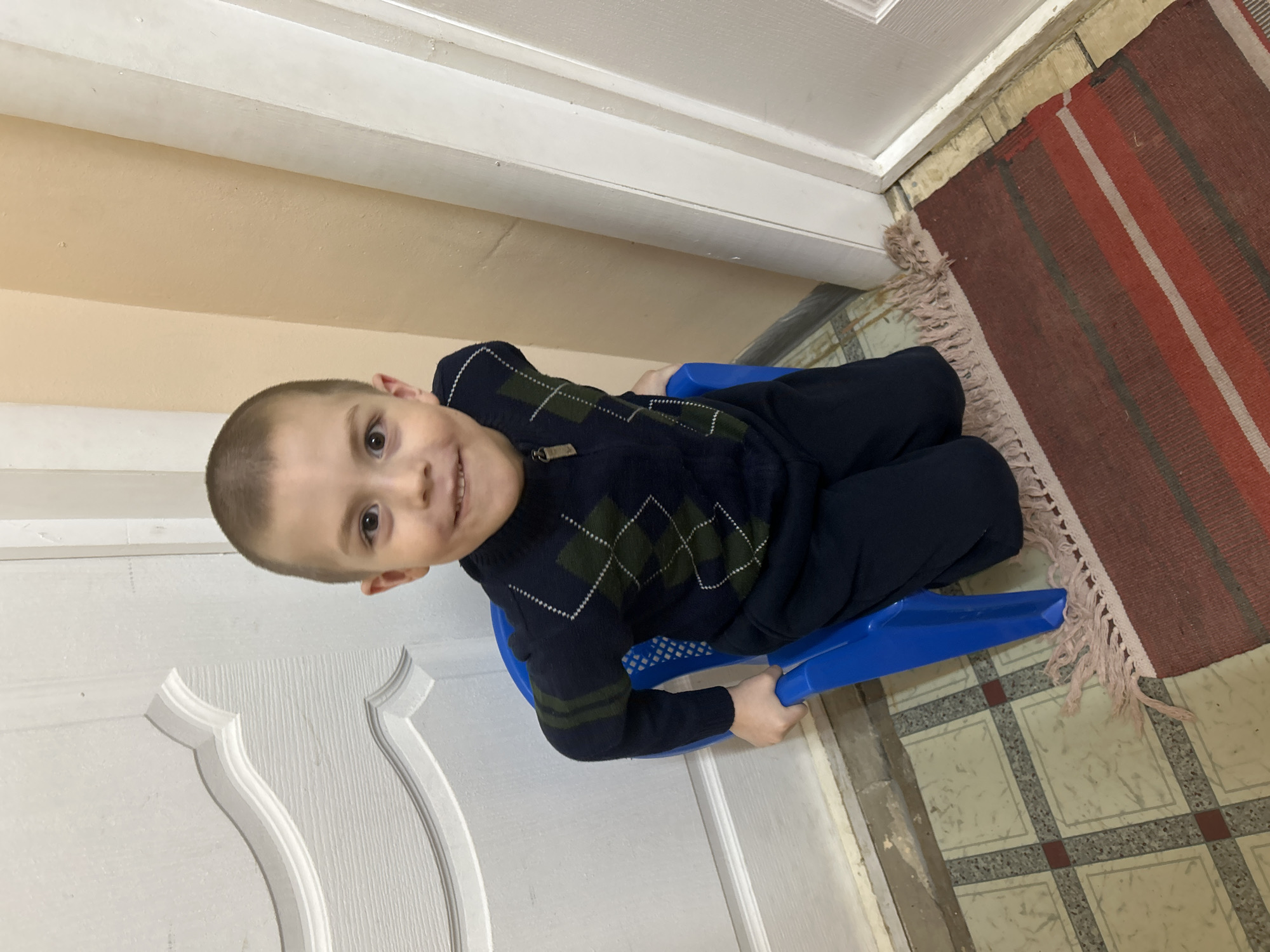 A young boy sits on a blue plastic sled indoors. He wears a dark blue sweater with a green diamond pattern and dark pants. The sled is positioned near a white door with decorative molding. A colorful striped rug lies on a tiled floor with a square pattern. The wall is a light peach color. The image was captured indoors, likely in a home. There is no indication that the photo was taken in the city of Фоки, Russia.