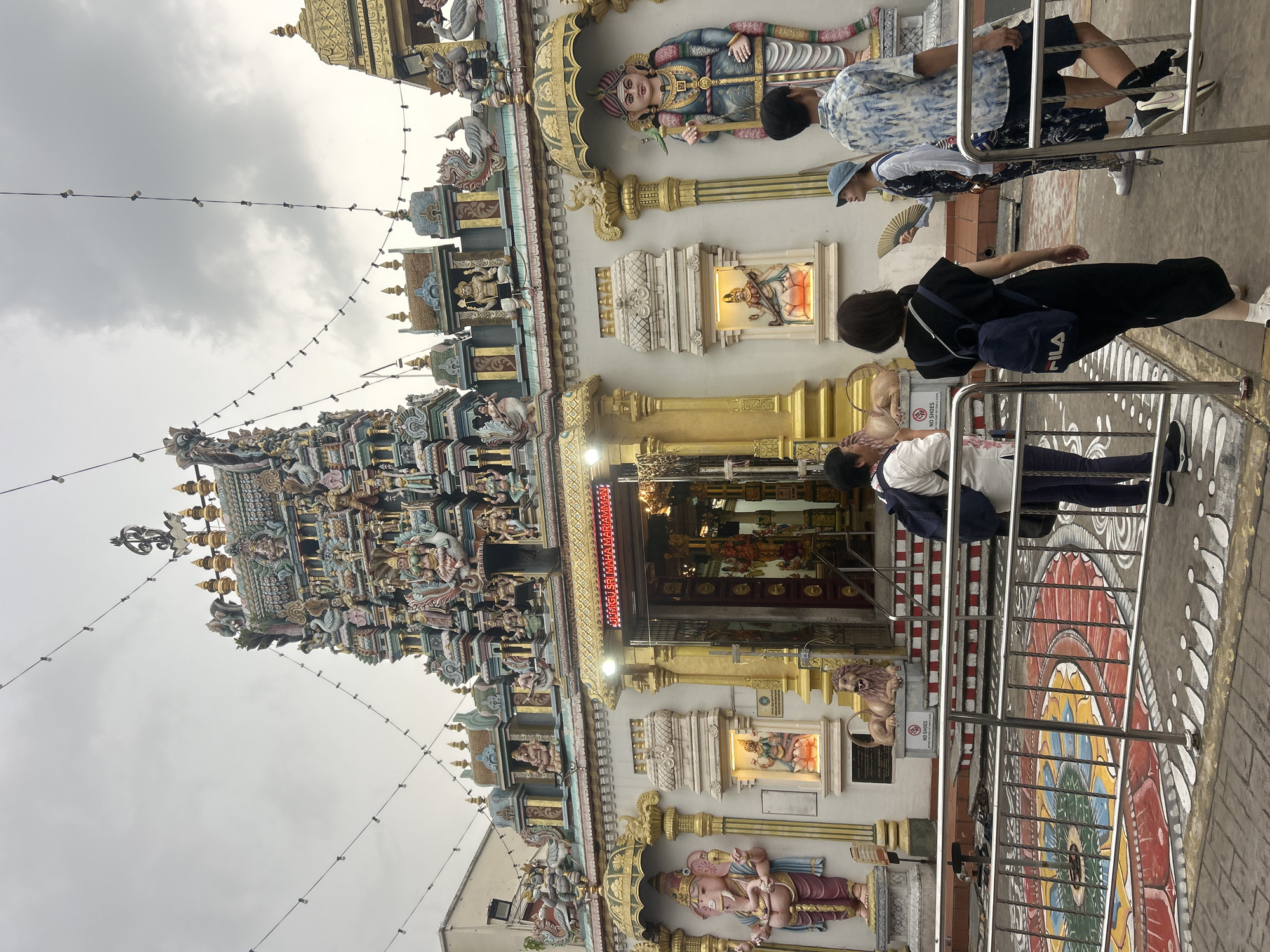This image depicts the Sri Maha Mariamman Temple, a prominent Hindu temple located in George Town, Penang, Malaysia. The temple's facade is elaborately decorated with colorful statues and intricate carvings, typical of South Indian architecture. In the foreground, a group of people are standing near the entrance, some are looking at the temple, others are walking. They appear to be tourists or devotees visiting the temple. The area in front of the temple features a patterned floor design and a metal railing. The sky is overcast, suggesting a cloudy day. The overall scene conveys a sense of cultural and religious significance.