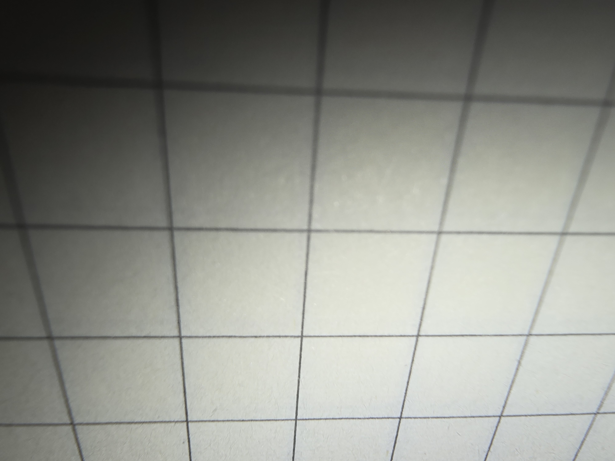 The image shows a close-up, macro view of a grid-lined paper surface, likely from a notebook. The lines are thin and dark, forming squares. The paper itself appears to have a slight texture. The lighting creates a gradient, with the top left area being brighter and the bottom right area becoming progressively darker and more shadowed. There are no people, subjects, objects, or actions depicted. The image does not contain any information that would allow for its localization in Ulan-Ude, Russia, or any other specific geographical location.